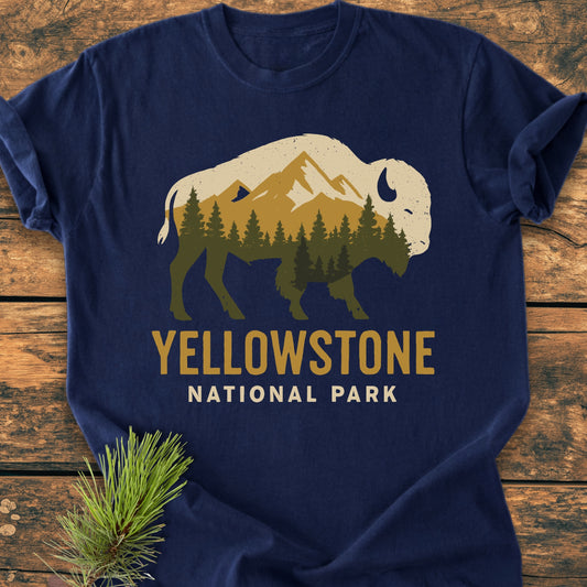 Yellowstone Bison - Vintage Wash Tee features a navy-blue color with a bison graphic filled with mountains and pine trees, and YELLOWSTONE NATIONAL PARK text. Laid on wood, adorned with a pine sprig.