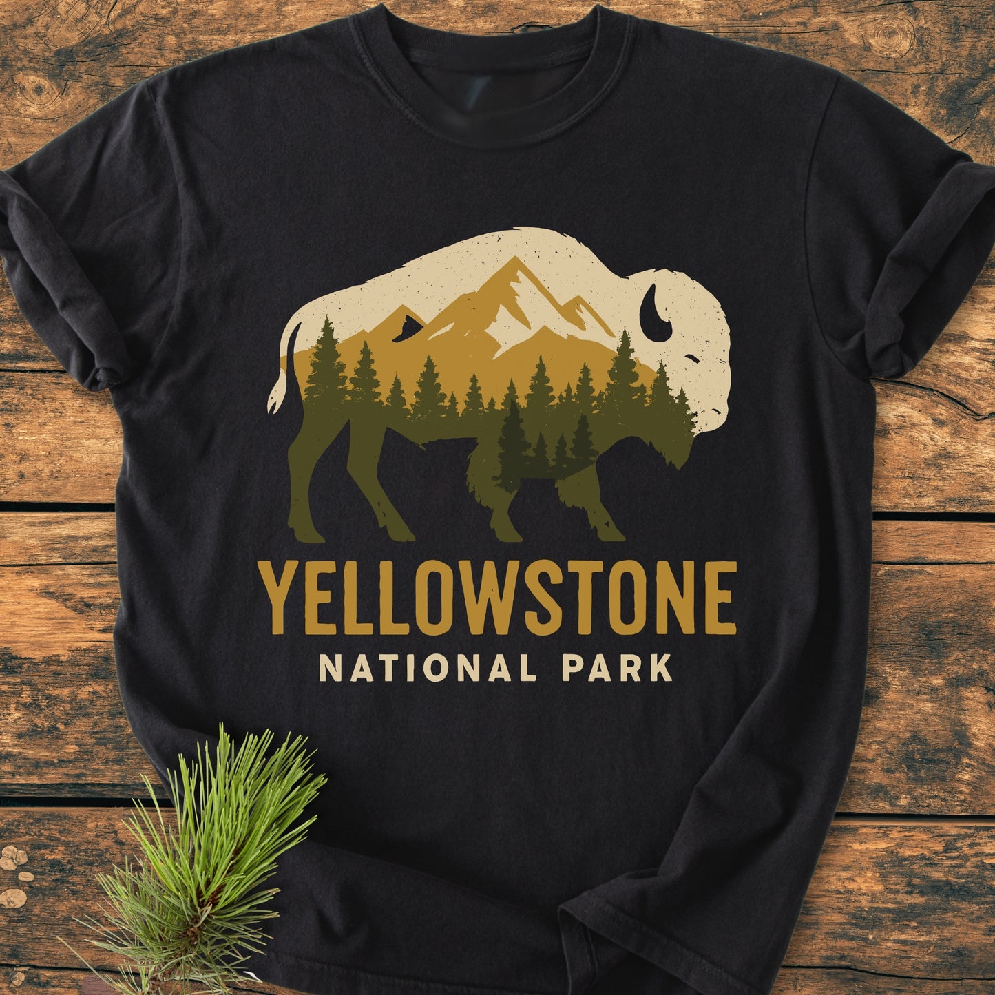 Yellowstone Bison - Vintage Wash Tee features a bison silhouette with a mountain scene. YELLOWSTONE NATIONAL PARK is printed below. Displayed on a wooden surface with pine needles accentuating its rustic appeal.