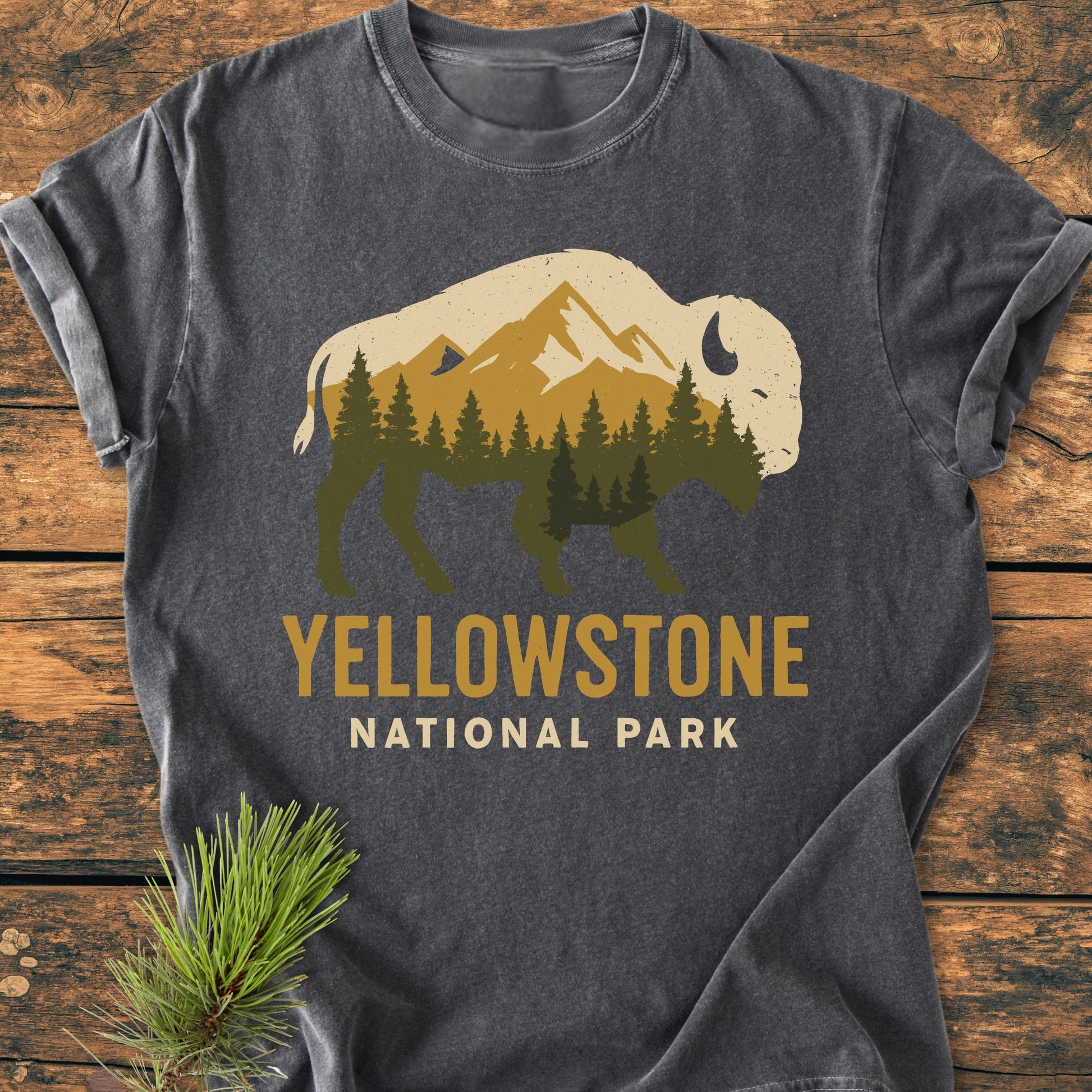 Yellowstone Bison - Vintage Wash Tee features a bison silhouette with mountains and forest print, laid flat on wood, with YELLOWSTONE NATIONAL PARK text. Accompanied by a pine branch for a rustic touch.
