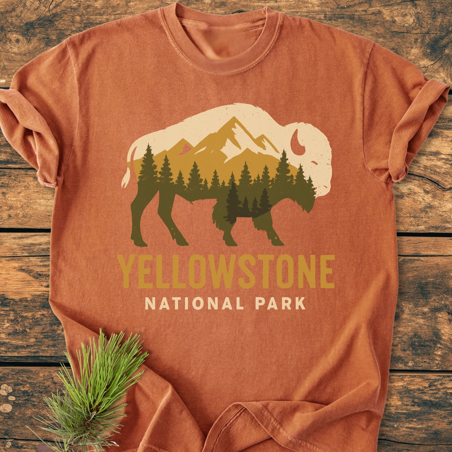 Yellowstone Bison - Vintage Wash Tee features a buffalo graphic filled with forest and mountains, “YELLOWSTONE NATIONAL PARK” text, laid on wood with a pine sprig beside it.