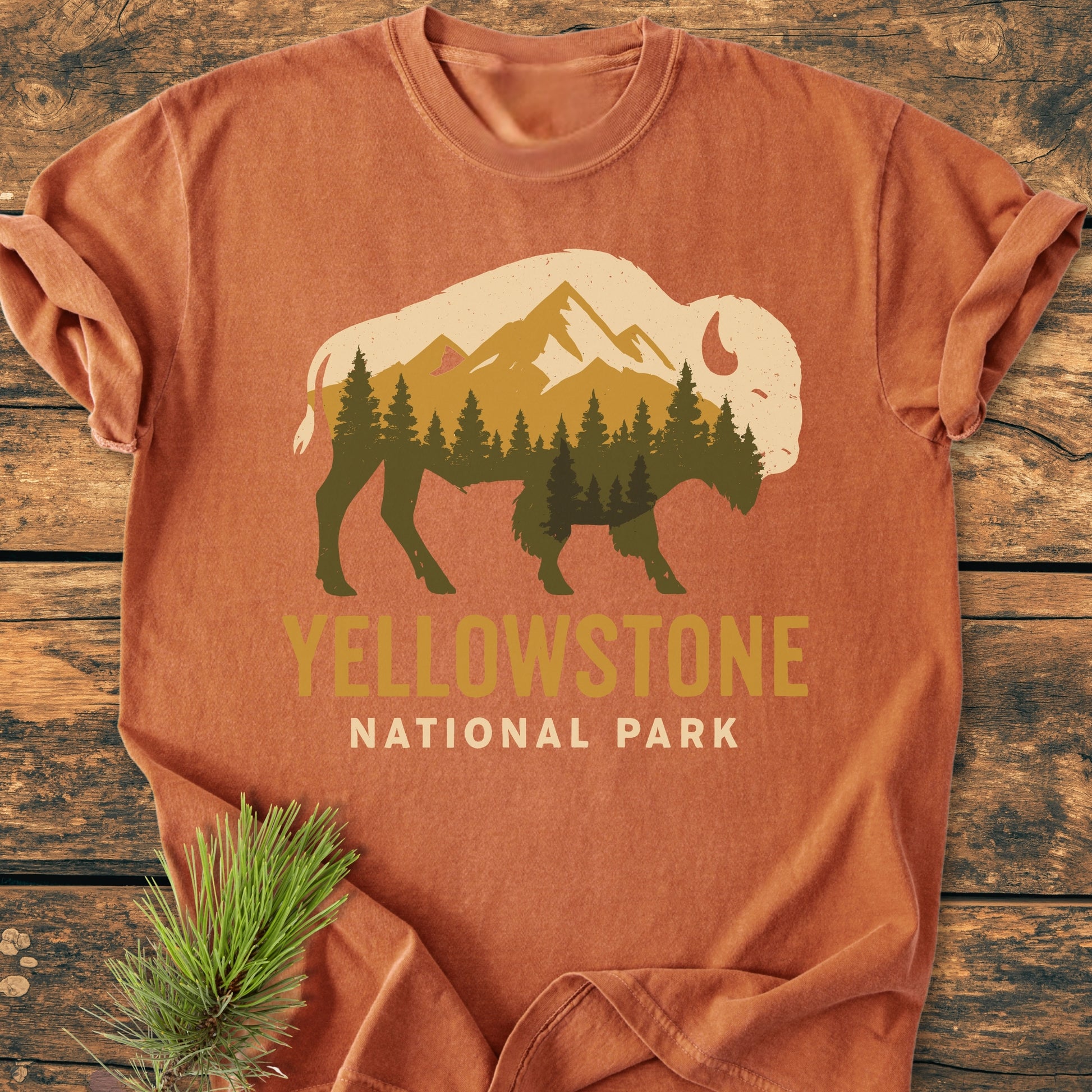 Yellowstone Bison - Vintage Wash Tee features a buffalo graphic filled with forest and mountains, “YELLOWSTONE NATIONAL PARK” text, laid on wood with a pine sprig beside it.