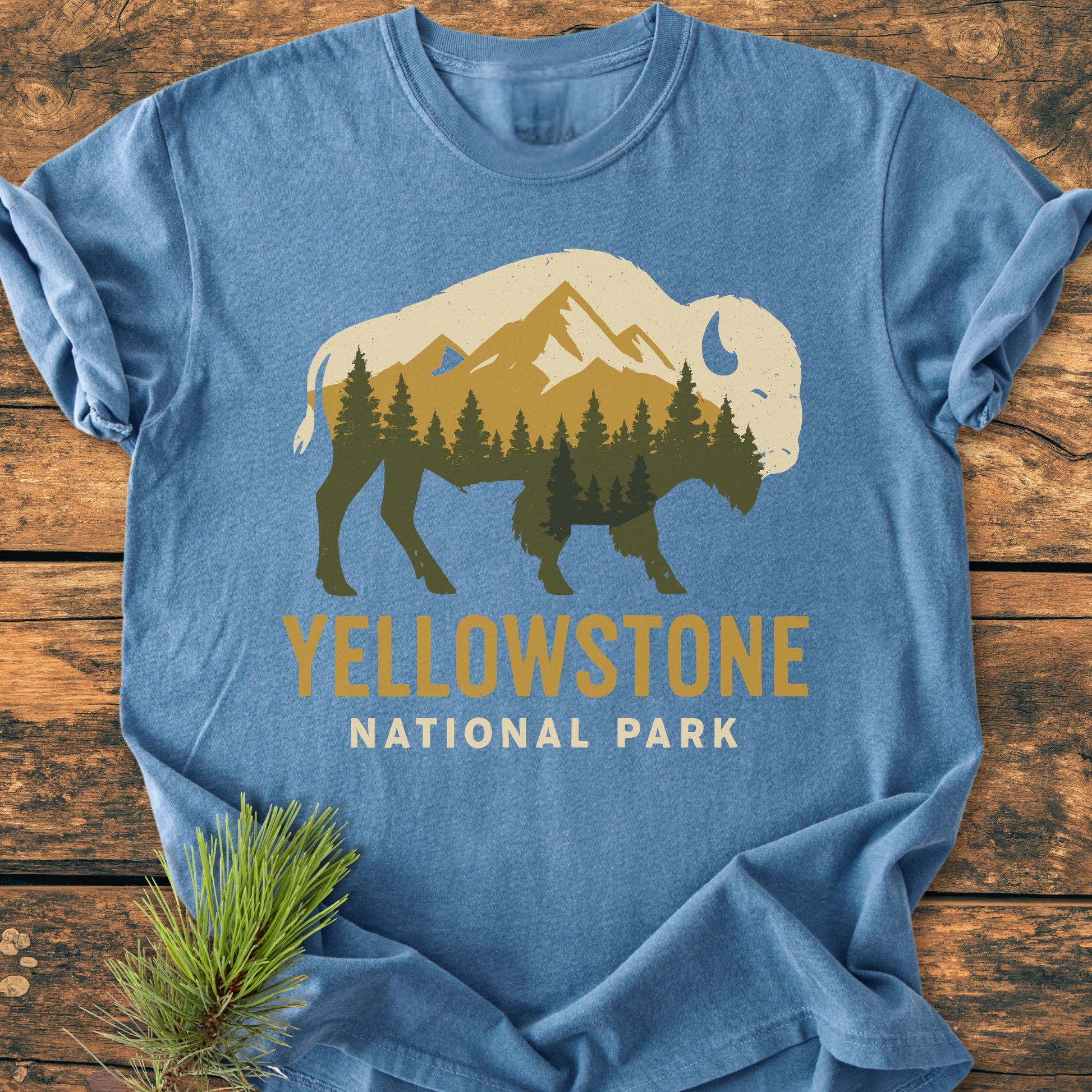 Yellowstone Bison - Vintage Wash Tee featuring a bison silhouette with mountain scenery, laid on wood. YELLOWSTONE NATIONAL PARK text below. Pine sprig beside the 100% ring-spun cotton garment.