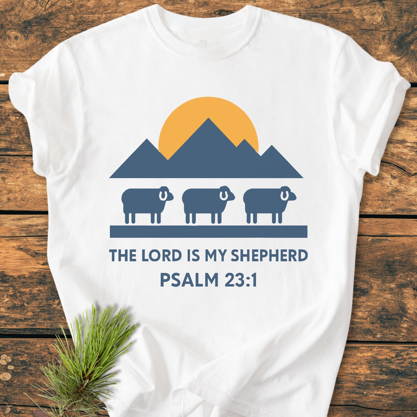 The Lord Is My Shepherd - Vintage Wash Tee features a mountain and sun graphic with sheep silhouettes and Psalm 23:1 text, laid flat on a wooden surface, showcasing its faith-based design.