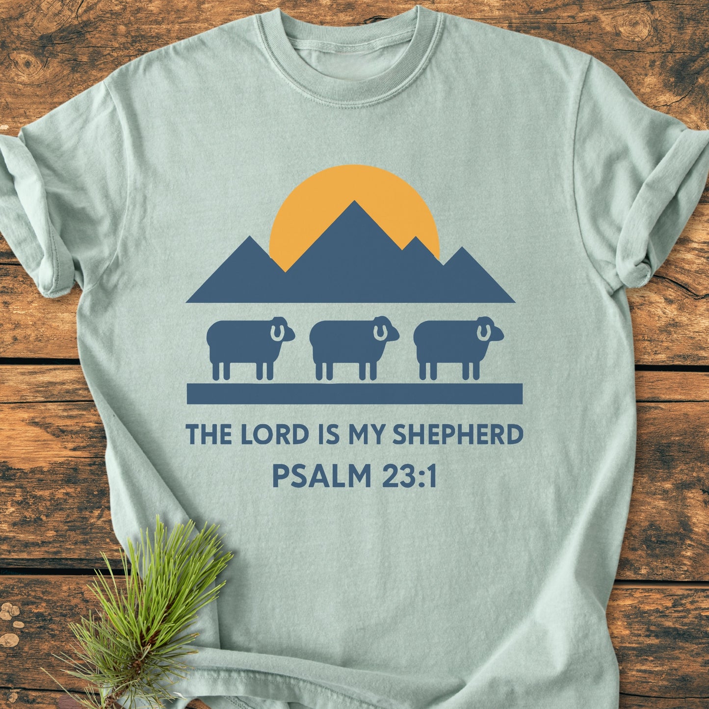 The Lord Is My Shepherd - Vintage Wash Tee features a sunrise over mountains with sheep silhouettes and Psalm 23:1 text, laid on wood with a pine sprig near the hem.