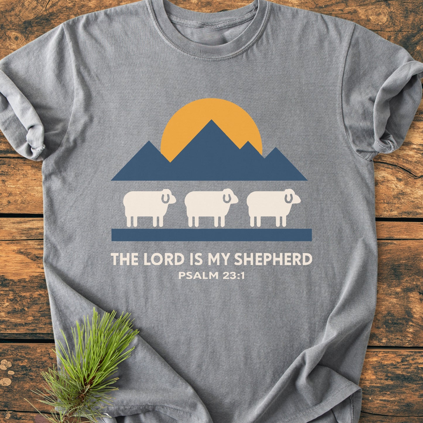 The Lord Is My Shepherd - Vintage Wash Tee features a mountain and sheep graphic with Psalm 23:1 text on a heather-gray T-shirt laid on wooden planks, accented by a pine sprig.