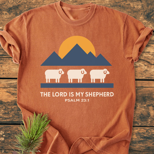 The Lord Is My Shepherd - Vintage Wash Tee displays a sun, mountains, and sheep graphic, with scripture text, on a rust-orange T-shirt, laid on wood with a pine sprig beside it.
