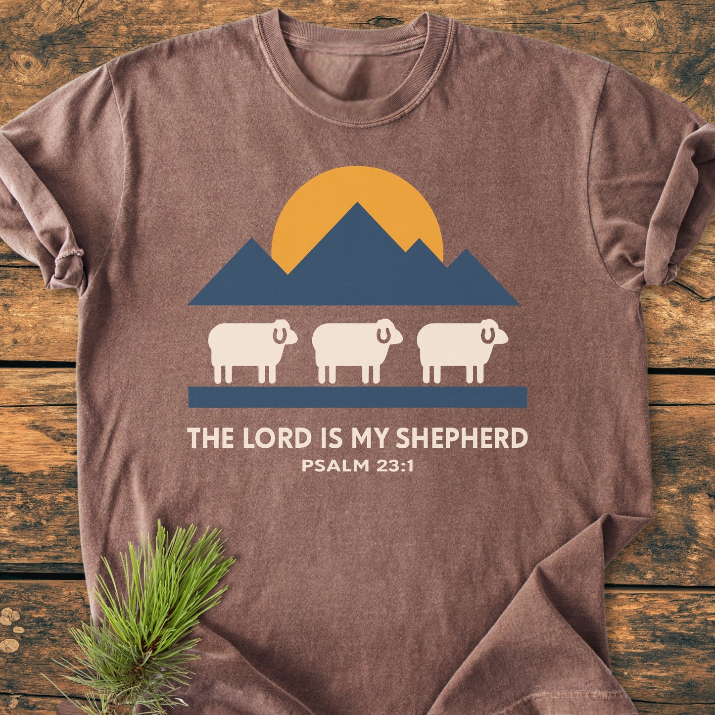 The Lord Is My Shepherd - Vintage Wash Tee features a mountain graphic, sun, and sheep with Psalm 23:1 text on a brown Comfort Colors t-shirt, laid on a wooden surface.