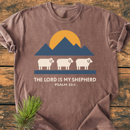 The Lord Is My Shepherd - Vintage Wash Tee features a mountain graphic, sun, and sheep with Psalm 23:1 text on a brown Comfort Colors t-shirt, laid on a wooden surface.