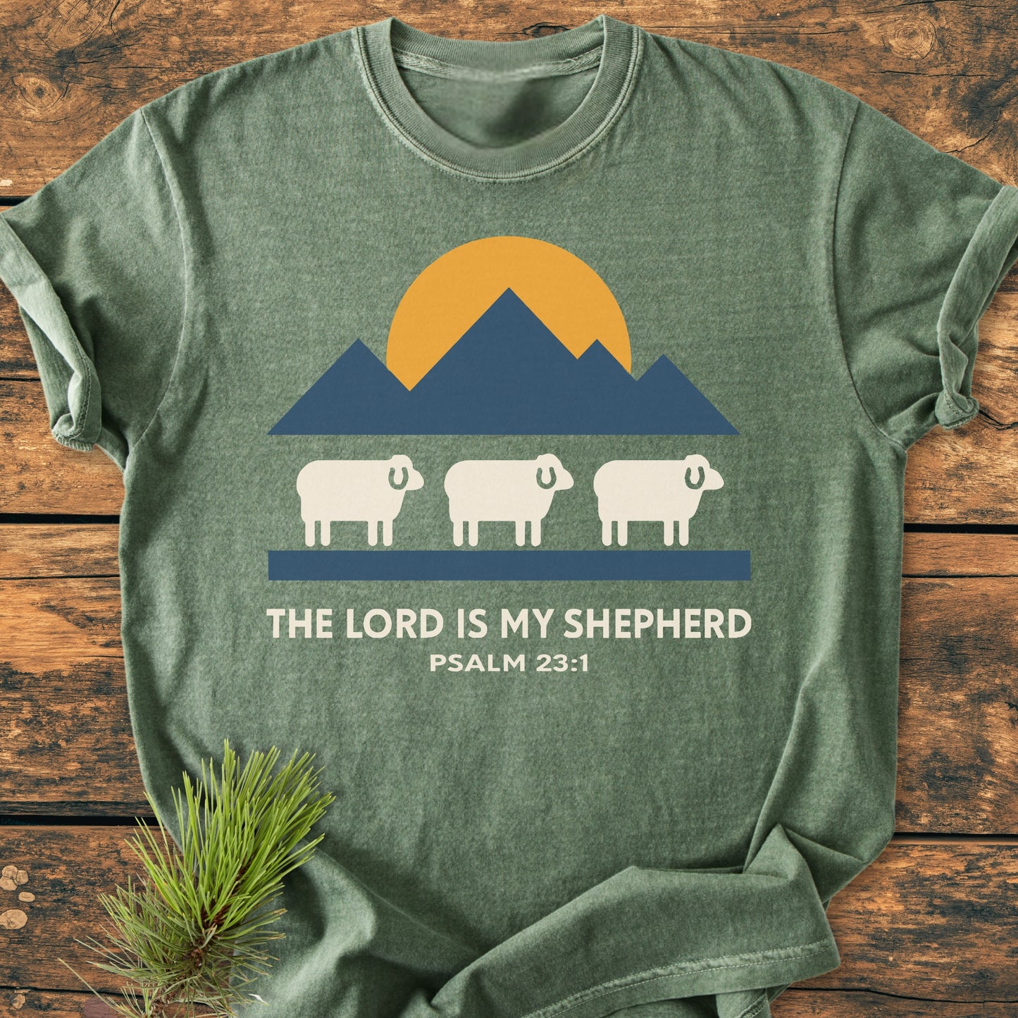 The Lord Is My Shepherd - Vintage Wash Tee featuring a mountain graphic and sheep with Psalm 23:1 text, laid on wood with pine sprig, highlighting its relaxed fit and soft-washed texture.