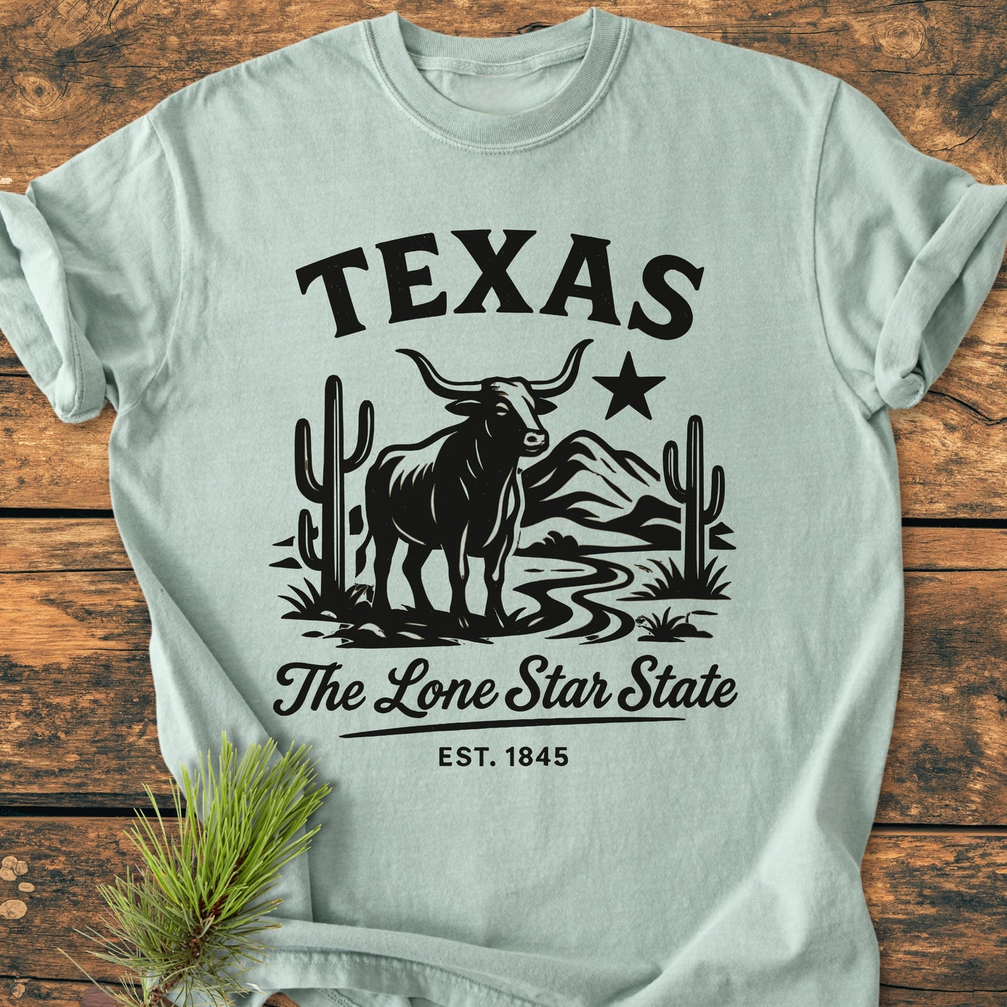 Texas, The Lone Star State - Vintage Wash Tee featuring bold TEXAS text, longhorn steer graphic, and The Lone Star State script, displayed on a wooden surface with a pine sprig accent.