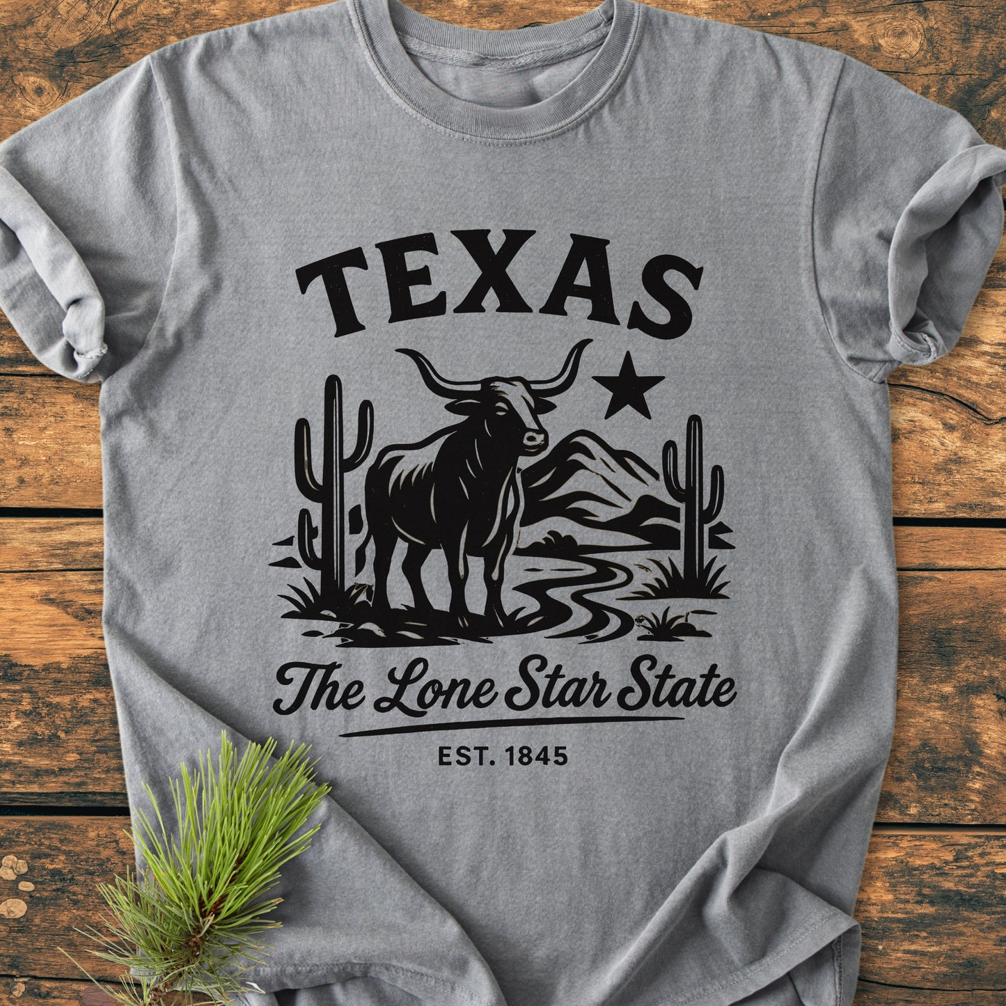 Texas, The Lone Star State - Vintage Wash Tee features a black graphic of a long-horn steer, cactus, mountain scene, and text on a heather-gray background, laid flat with pine sprig accent.