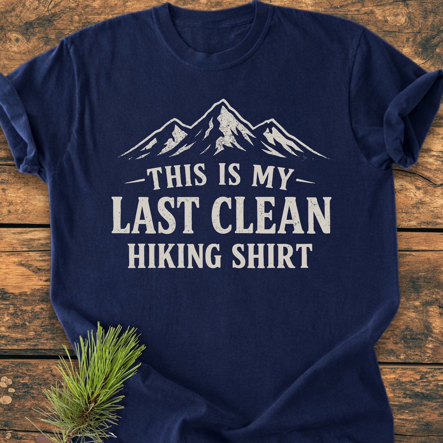 Navy-blue Last Clean Hiking Shirt - Vintage Wash Tee with a stylized mountain graphic and slogan, laid on wood with pine sprigs, showcasing its relaxed fit and durable, soft-washed fabric.