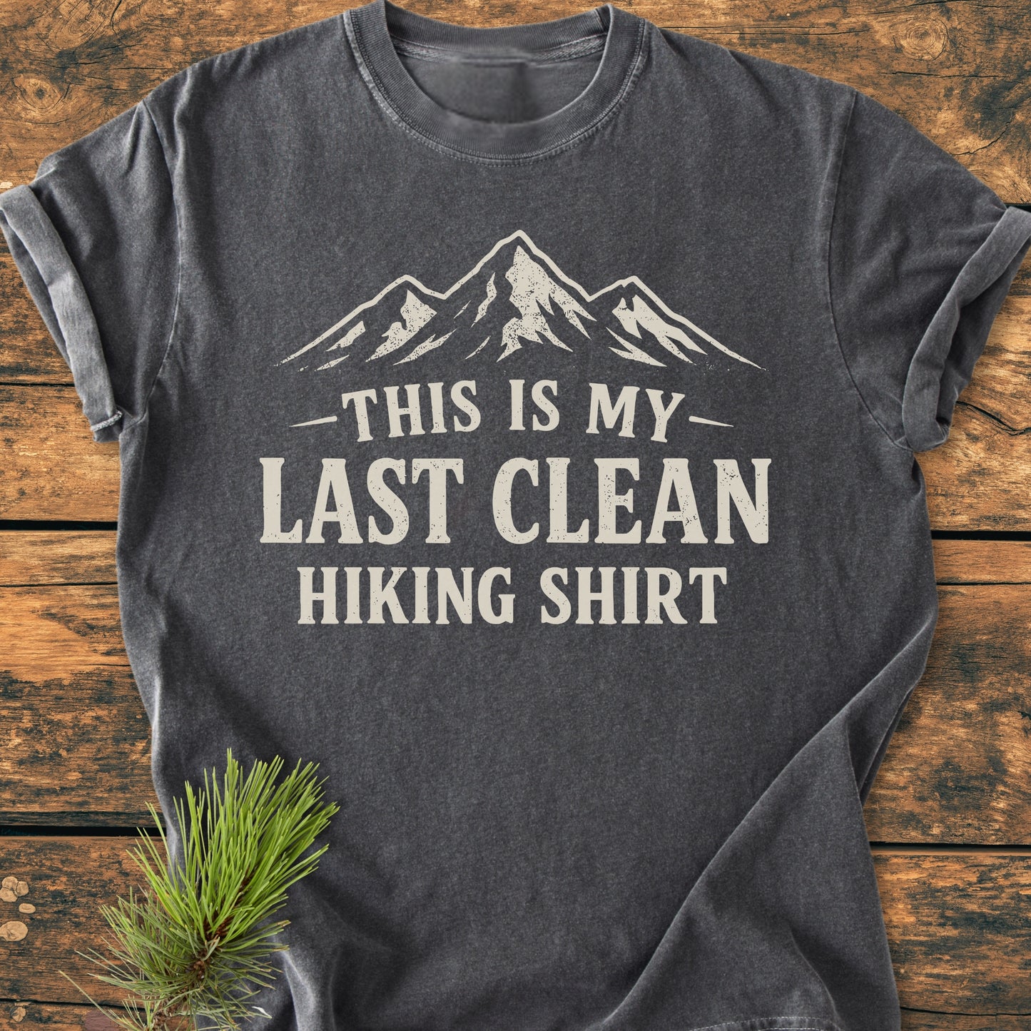 Charcoal-grey Last Clean Hiking Shirt - Vintage Wash Tee with mountain graphic, laid on wood, alongside a pine sprig. Made from soft, durable, 100% ring-spun cotton for ultimate comfort.