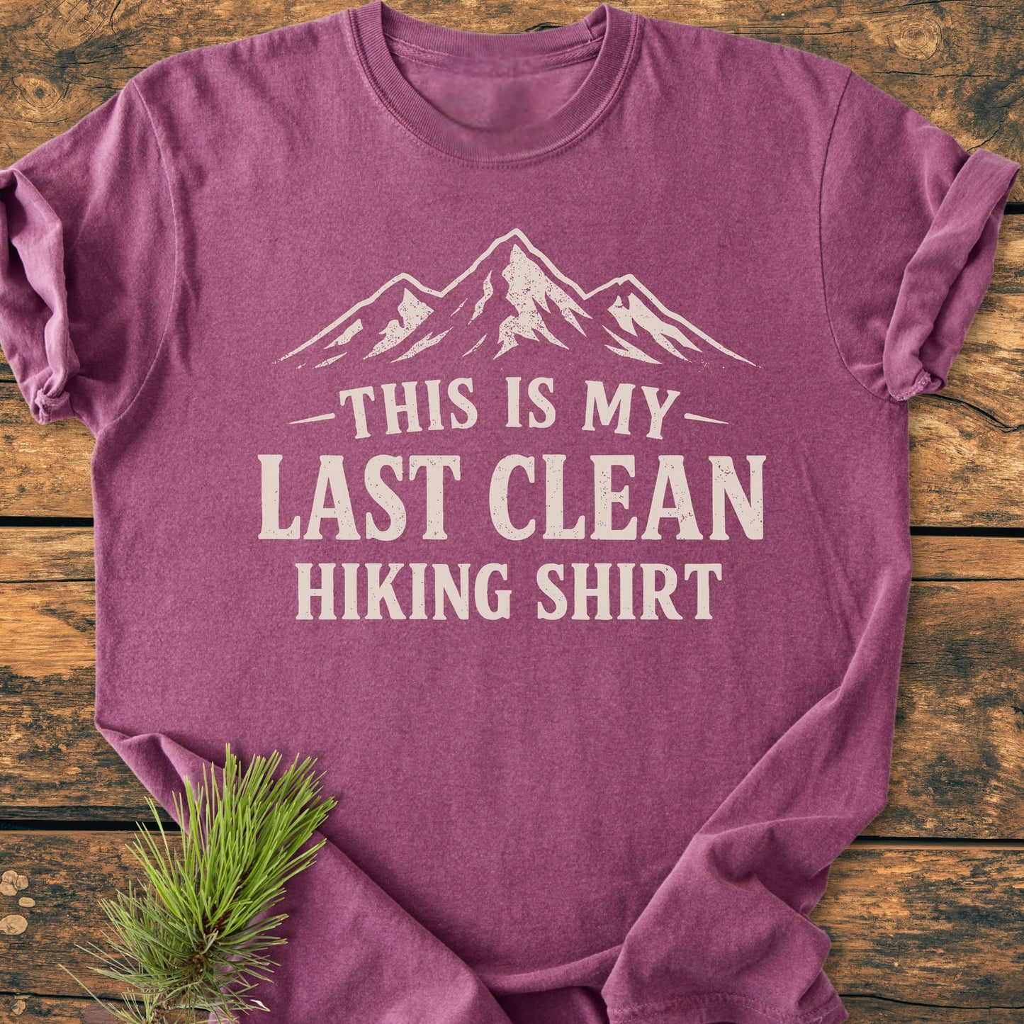 Last Clean Hiking Shirt - Vintage Wash Tee features a heather-purple crew-neck design with an off-white mountain graphic and slogan, styled on a wooden surface with a pine branch accent.