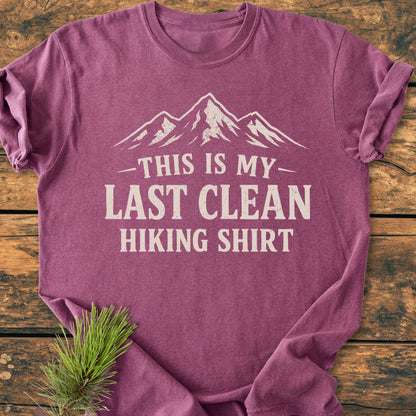 Last Clean Hiking Shirt - Vintage Wash Tee features a heather-purple crew-neck design with an off-white mountain graphic and slogan, styled on a wooden surface with a pine branch accent.