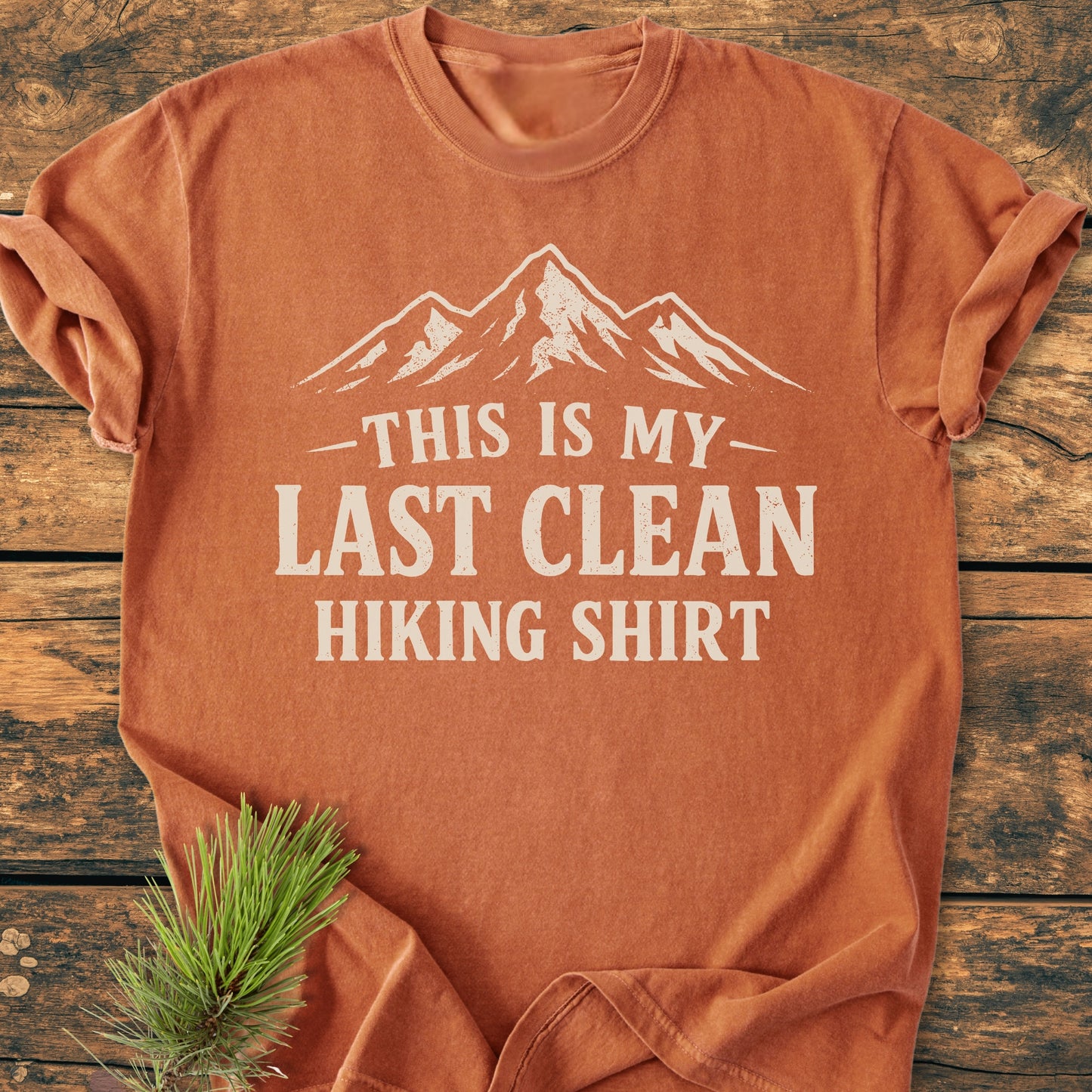 Last Clean Hiking Shirt - Vintage Wash Tee, featuring a mountain graphic and playful text, laid on wood with rolled sleeves and nearby pine sprig, showcasing its relaxed fit and durable stitching.