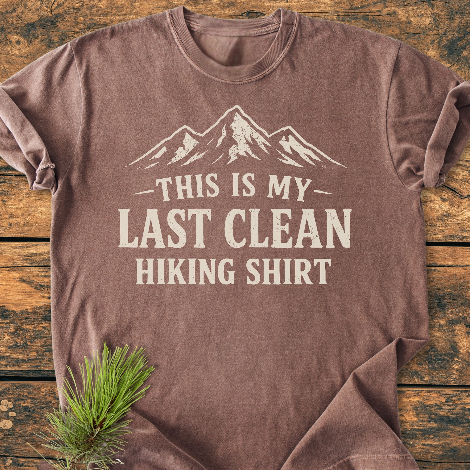 Last Clean Hiking Shirt - Vintage Wash Tee features a faded brown color with a white mountain graphic and bold text. Displayed on rustic wood, it embodies an outdoorsy theme with a pine sprig accent.