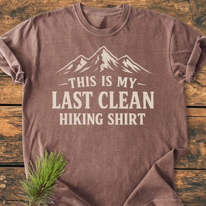 Last Clean Hiking Shirt - Vintage Wash Tee features a faded brown color with a white mountain graphic and bold text. Displayed on rustic wood, it embodies an outdoorsy theme with a pine sprig accent.