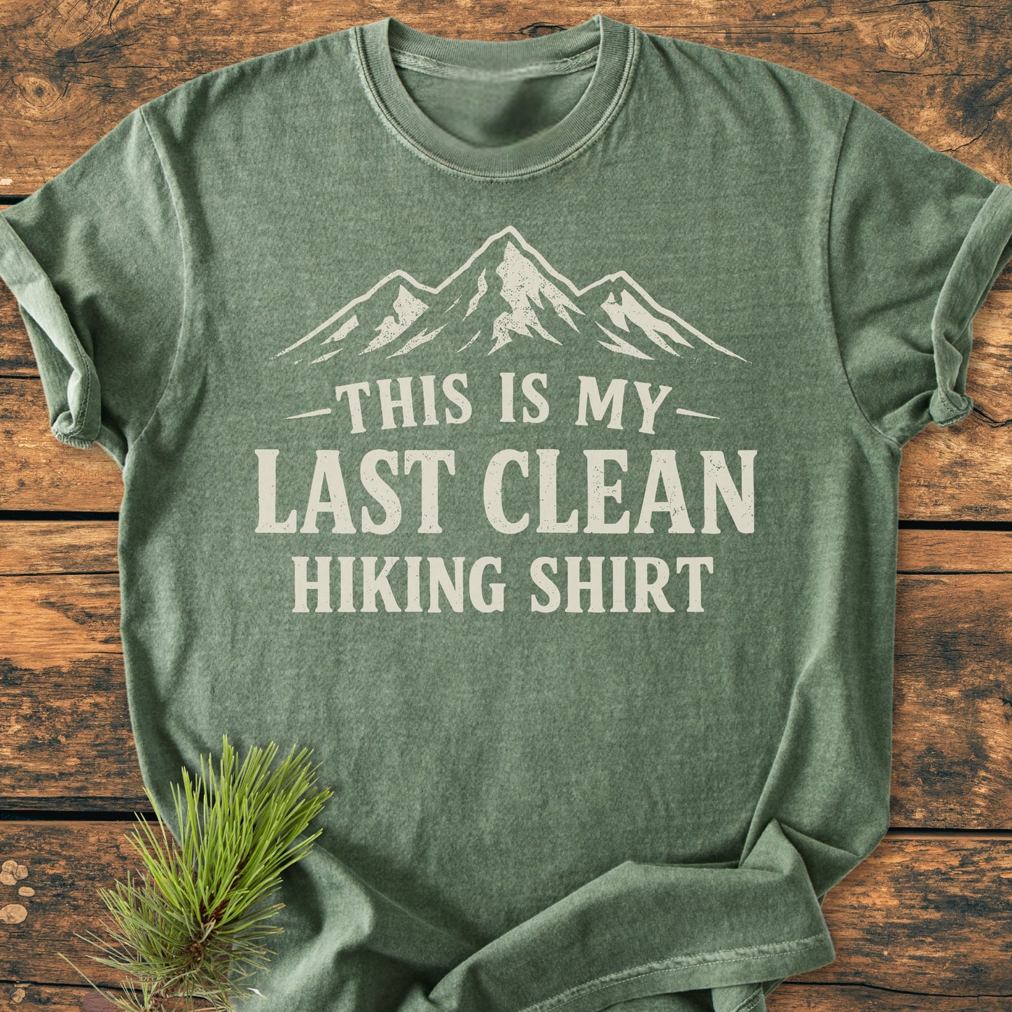 Last Clean Hiking Shirt - Vintage Wash Tee features a stylized mountain design with bold text, laid flat on wood, accented by a pine branch, highlighting its outdoor theme and relaxed fit.