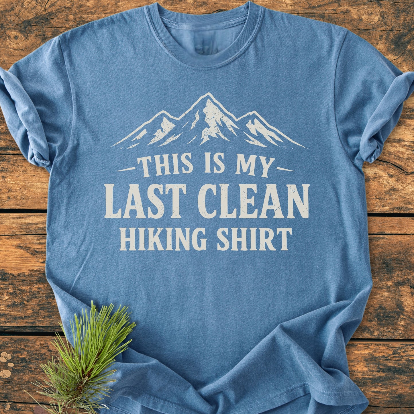 Heather-blue Last Clean Hiking Shirt - Vintage Wash Tee with white mountain graphic and bold text, laid flat on wood. Pine sprig accentuates the tee's rustic, outdoor theme.