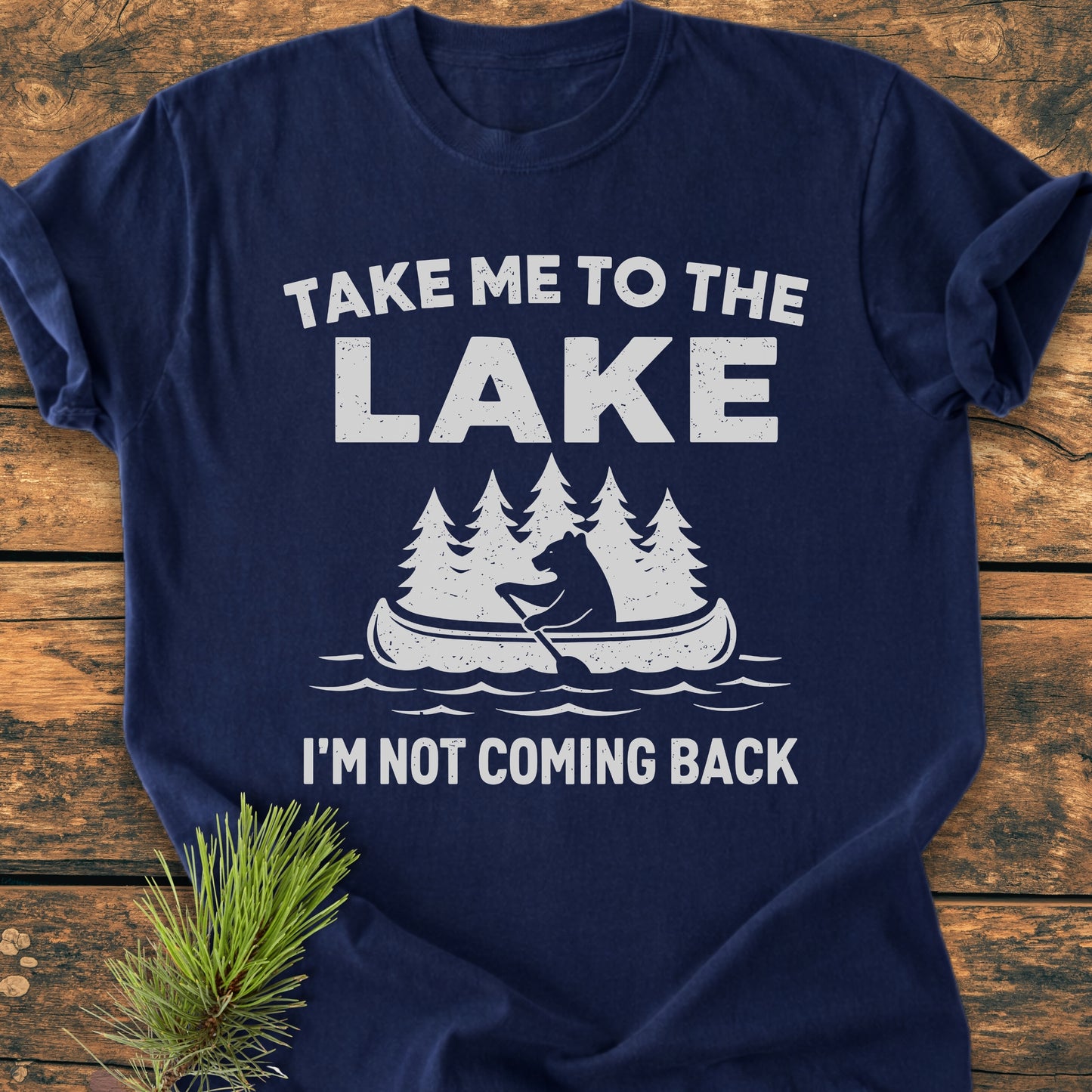 Take Me To The Lake - Vintage Wash Tee featuring a white design of a bear paddling a canoe, laid on a wooden surface with a pine sprig, showcasing its cozy and durable garment-dyed fabric.