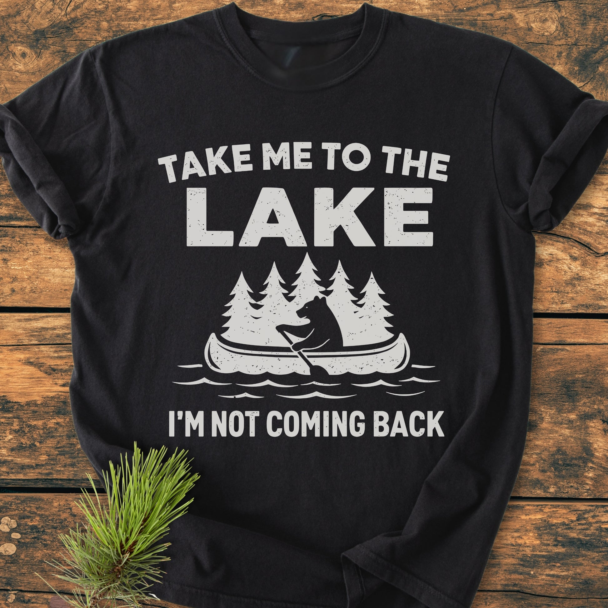 Take Me To The Lake - Vintage Wash Tee: Black T-shirt with white text and bear canoeing graphic, laid flat on wood. Features relaxed fit, garment-dyed 100% ring-spun cotton, emphasizing comfort and durability.