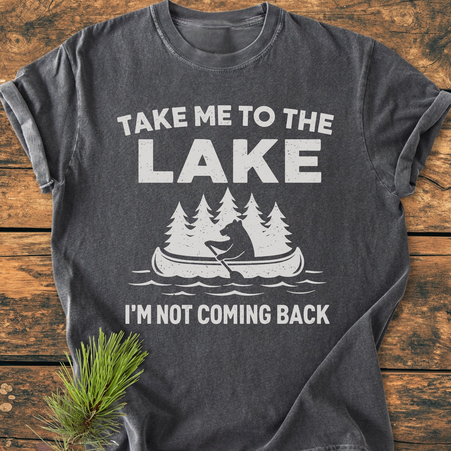 Take Me To The Lake - Vintage Wash Tee features a bear in a canoe with pine trees, printed on a charcoal-gray shirt. Laid on a wooden surface with a pine sprig beside it.