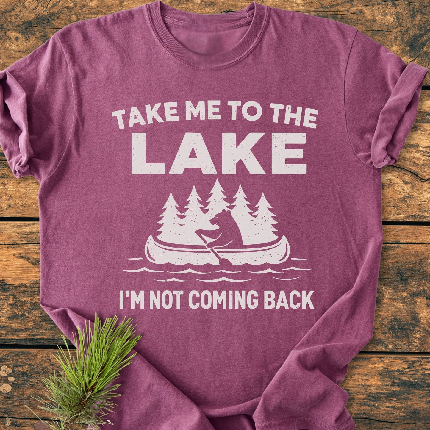 Take Me To The Lake - Vintage Wash Tee: Heather-purple short-sleeve shirt with bear canoe graphic, white text, laid on wood, accented by pine branch, embodying rustic charm and outdoor spirit.
