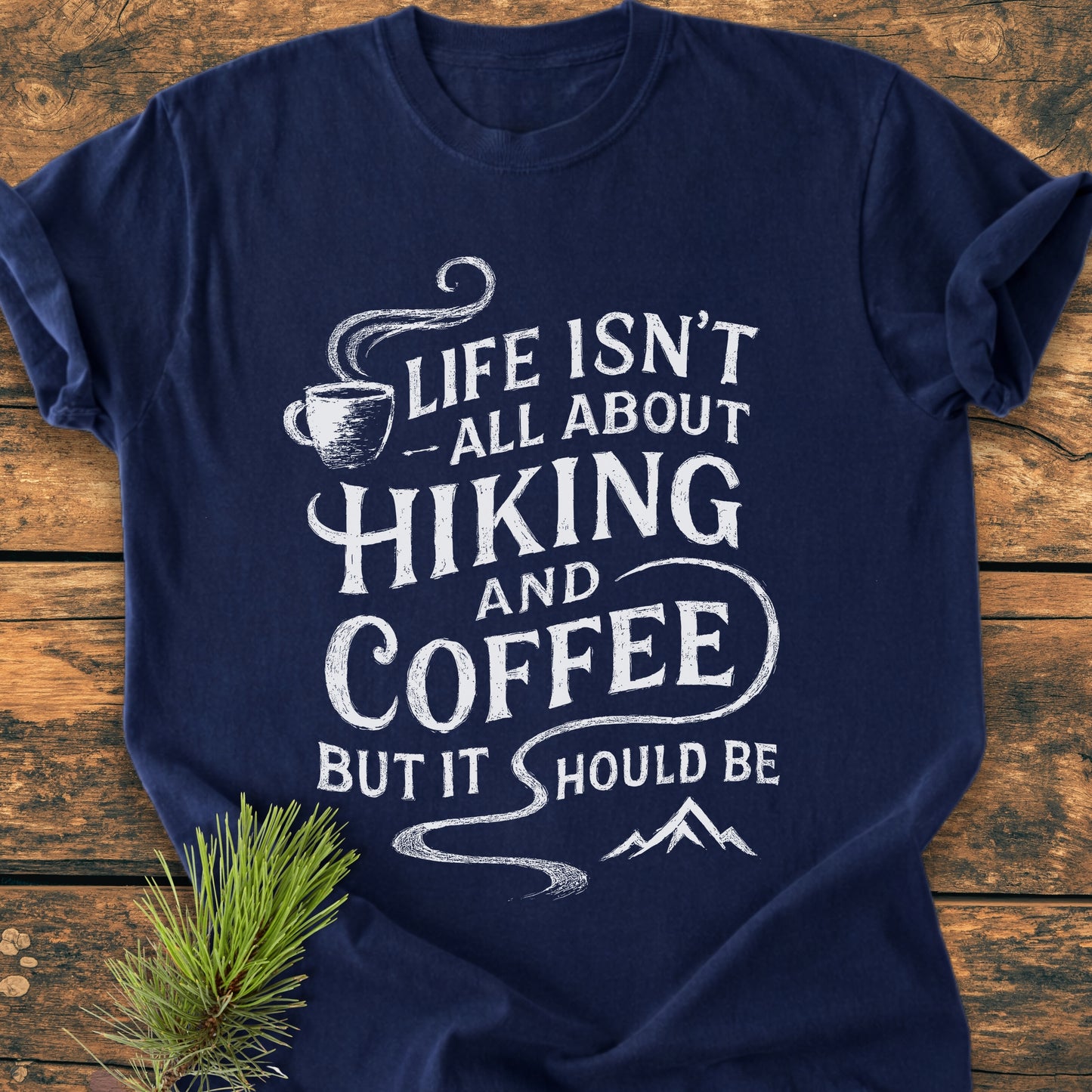 Hiking And Coffee - Vintage Wash Tee features a navy-blue design with a white slogan, coffee-cup icon, and mountain silhouette, emphasizing a rustic, hand-lettered style laid on a wooden surface.
