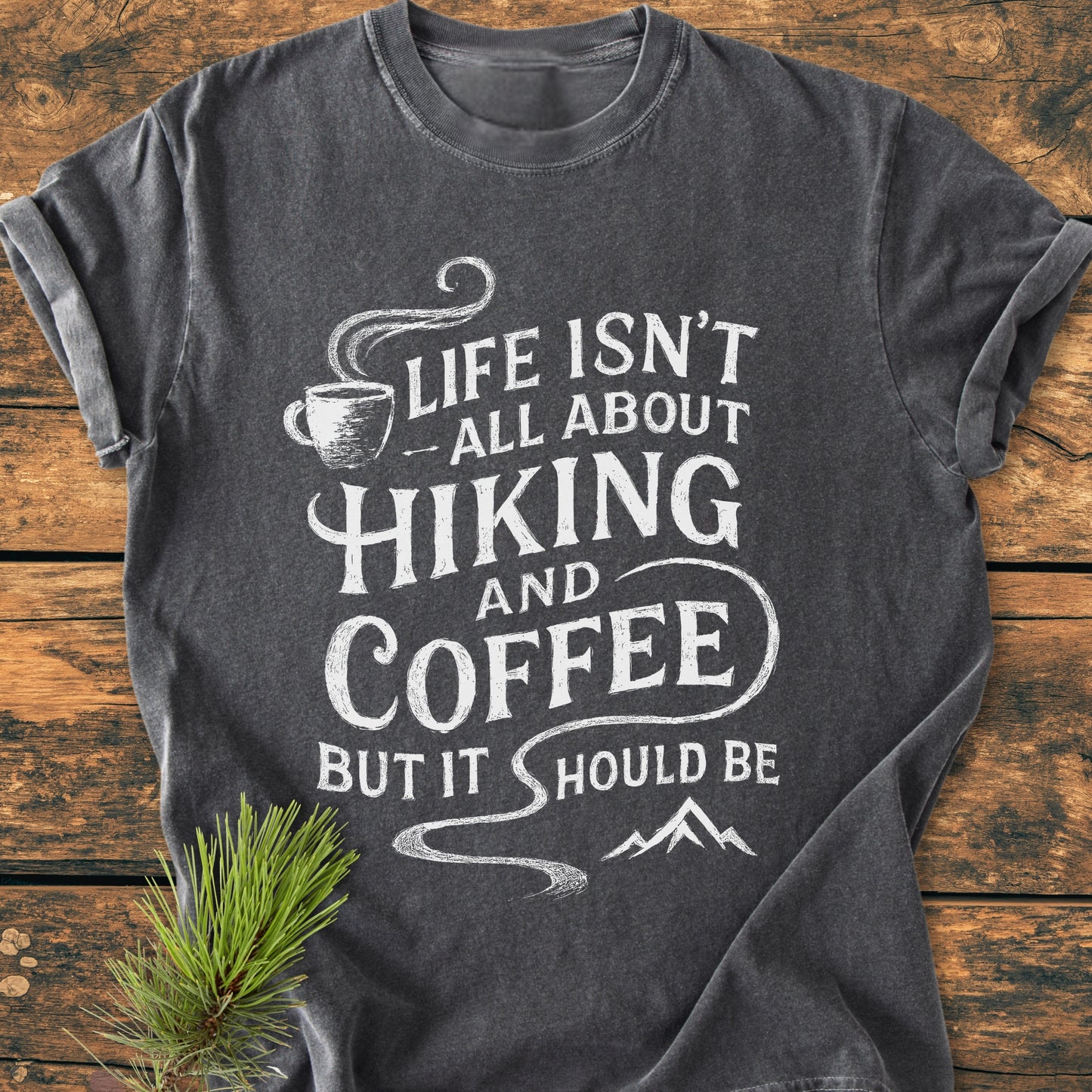 Hiking And Coffee - Vintage Wash Tee: Charcoal-gray tee with a hand-lettered hiking and coffee slogan, steaming cup, and mountain sketch, laid on wood, featuring a rustic pine branch accent.