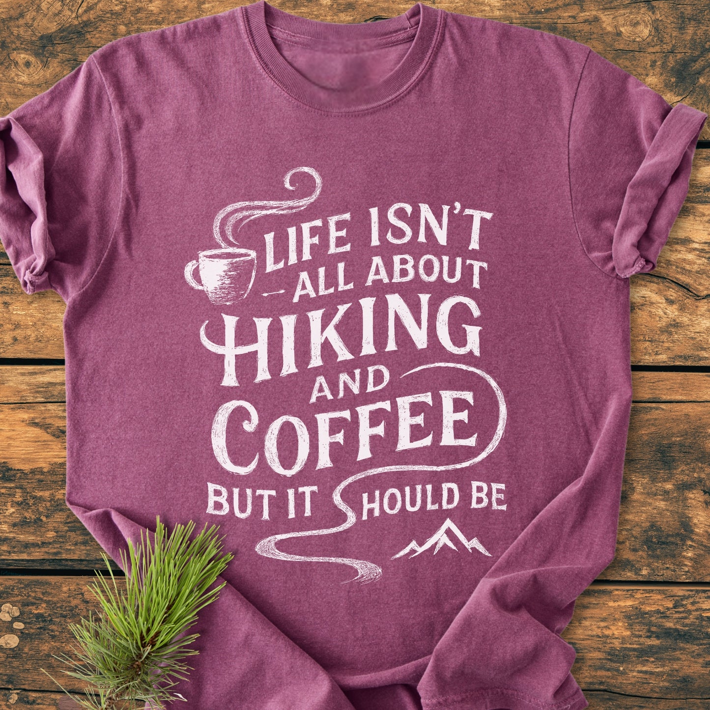Hiking And Coffee - Vintage Wash Tee features a white print of a steaming coffee cup and mountain on a mauve-purple background, laid flat on a wooden surface, with pine needles accenting.