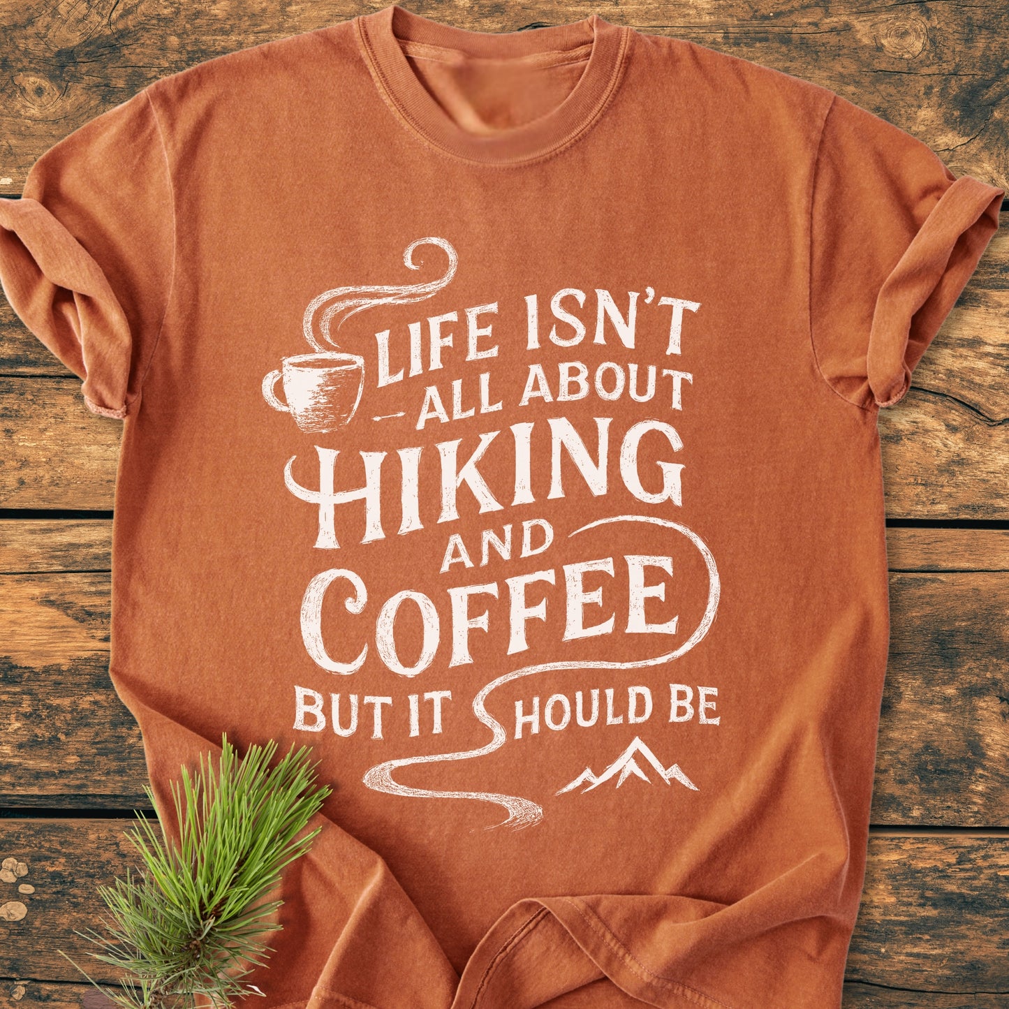 Hiking And Coffee - Vintage Wash Tee features a hand-lettered slogan and graphics of a coffee cup and mountains, laid on wood with a pine sprig, embodying an outdoorsy vibe.