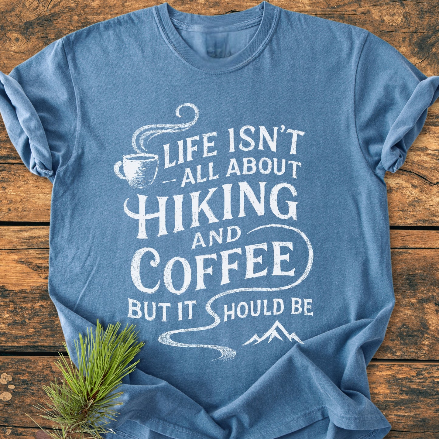 Hiking And Coffee - Vintage Wash Tee in heather-blue, featuring a white chalk-style slogan with a coffee cup and mountain graphics, laid flat on a wooden surface with a pine sprig accent.