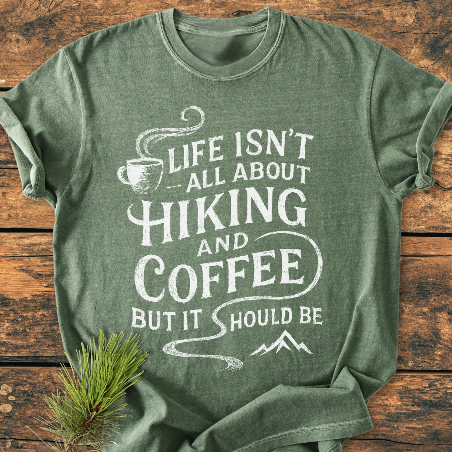 Hiking And Coffee - Vintage Wash Tee: Green T-shirt with a white design of a coffee cup, mountain, and slogan. Laid on wood with a pine sprig beside it, highlighting its cozy, relaxed fit.