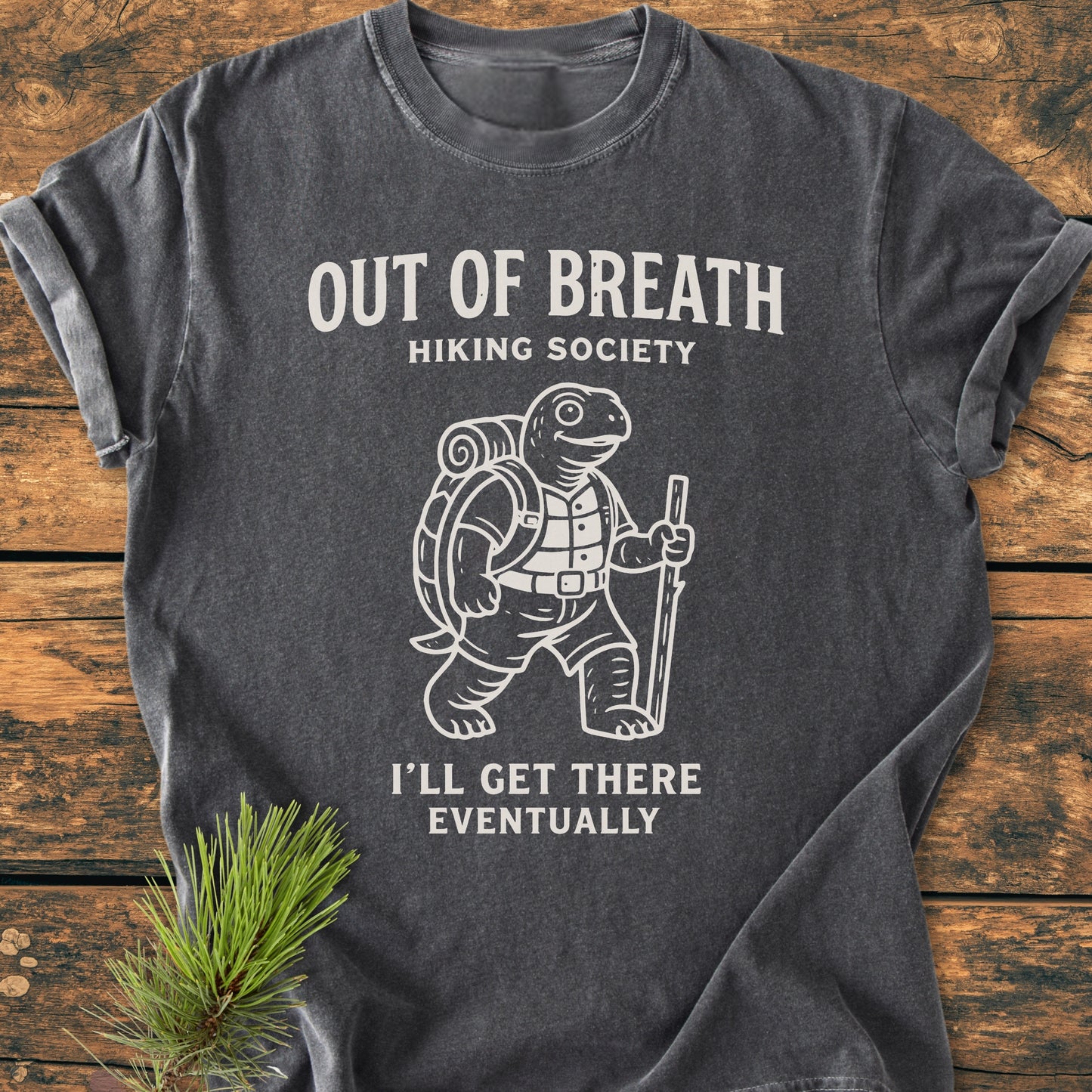 Charcoal-gray Out Of Breath Turtle - Vintage Wash Tee featuring a cartoon tortoise with hiking gear, screen-printed text, on a rustic surface with pine sprig accent.