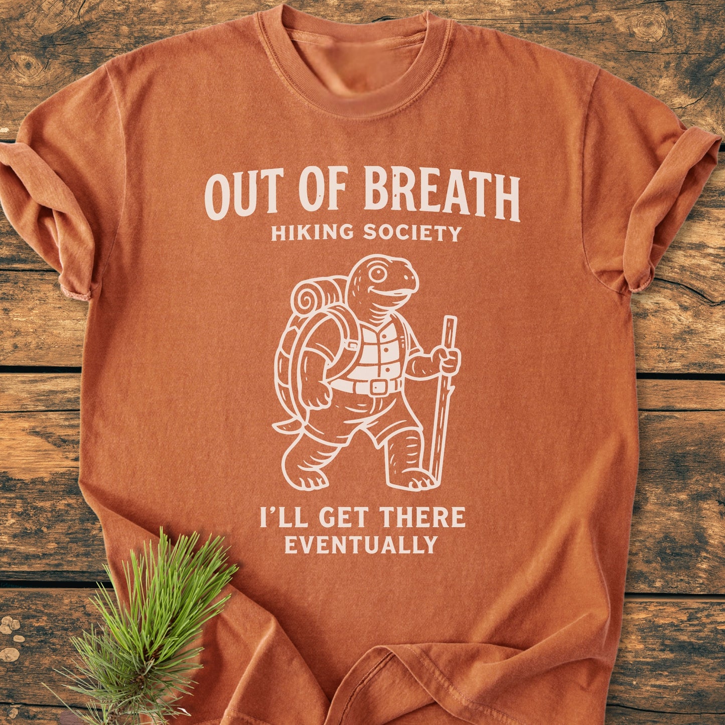 Out Of Breath Turtle - Vintage Wash Tee featuring a white line drawing of a turtle in hiking gear and humorous text, laid on wood with a pine sprig beside it.
