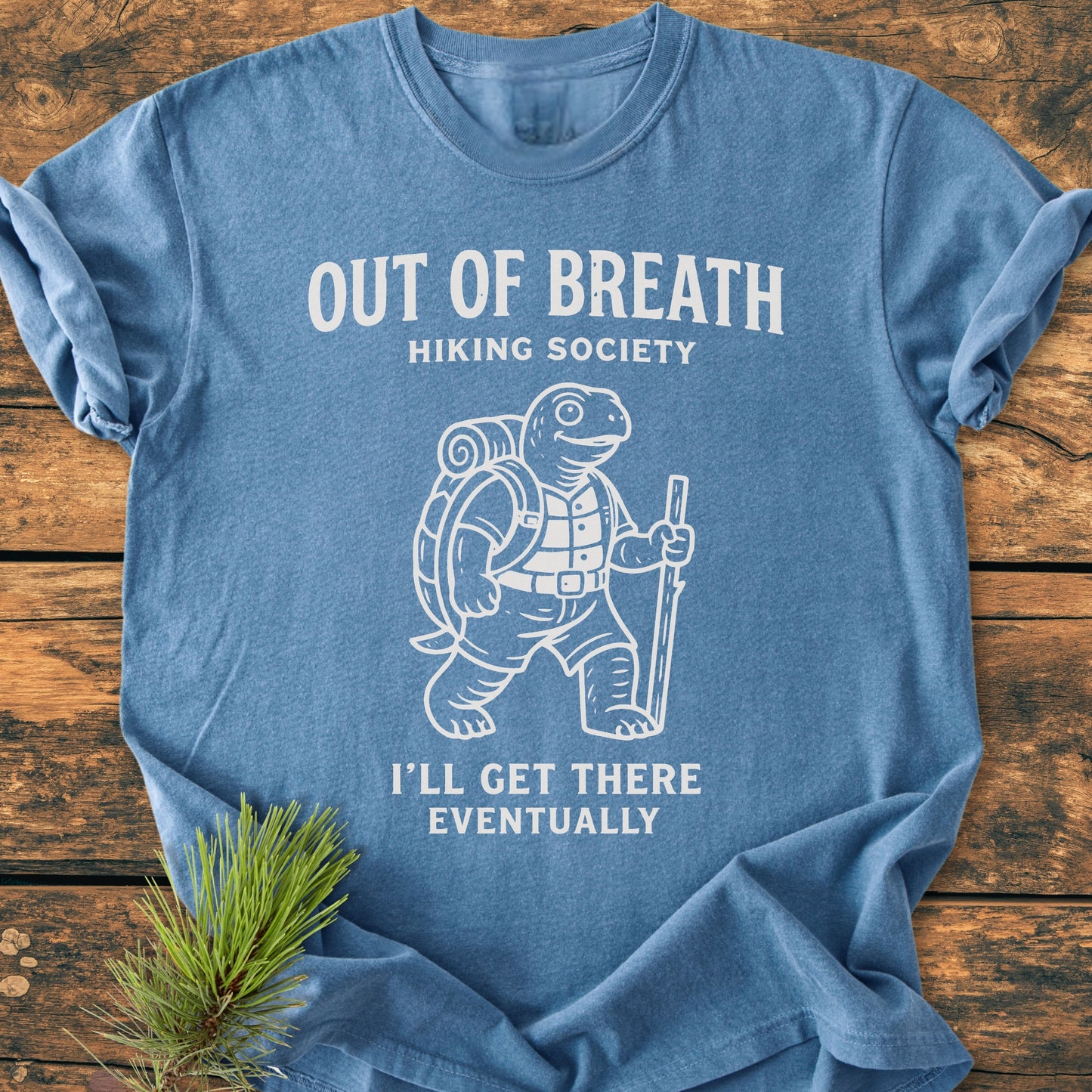 Light-blue Out Of Breath Turtle - Vintage Wash Tee featuring a cartoon tortoise with hiking gear, text above and below, laid on a wooden surface with a pine sprig beside it.