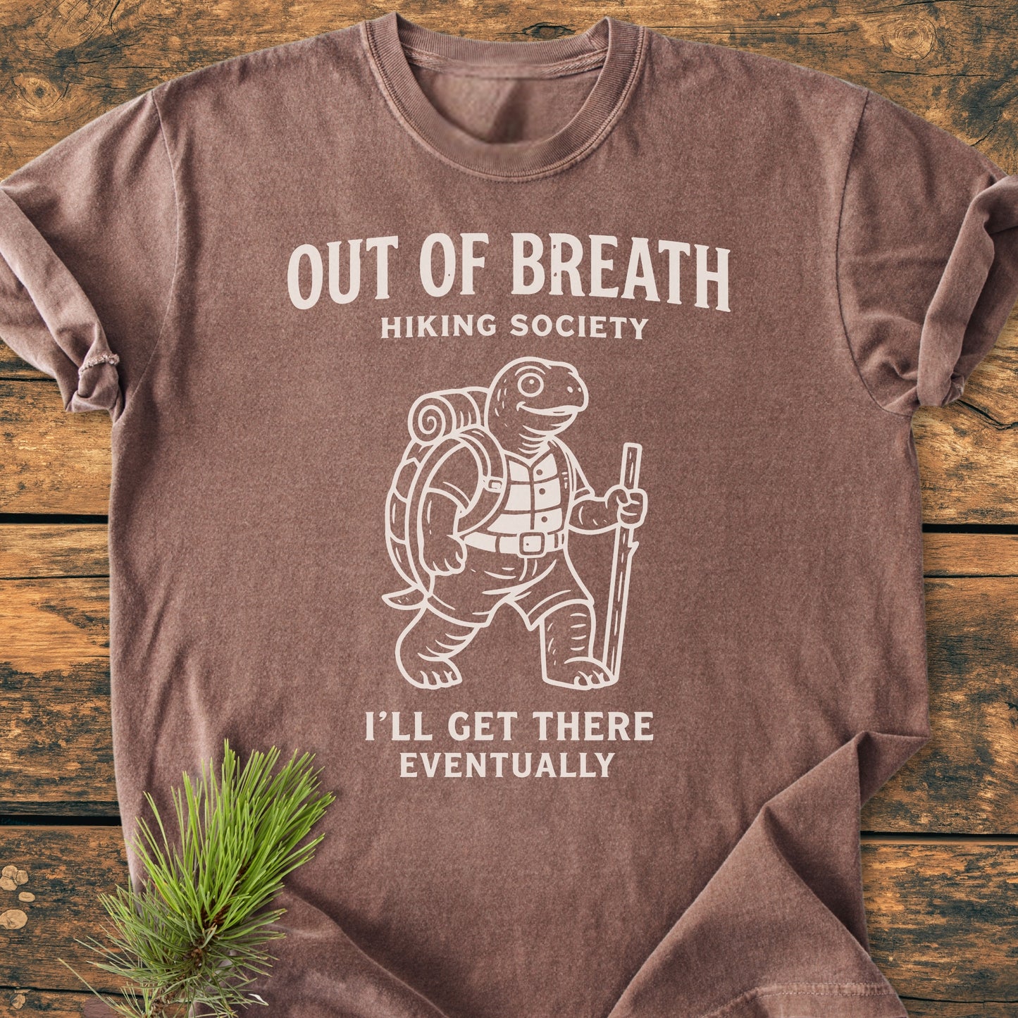 Out Of Breath Turtle - Vintage Wash Tee featuring a cartoon turtle hiking design and playful text, displayed on a wood surface with a pine sprig accentuating the shirt's relaxed, durable style.