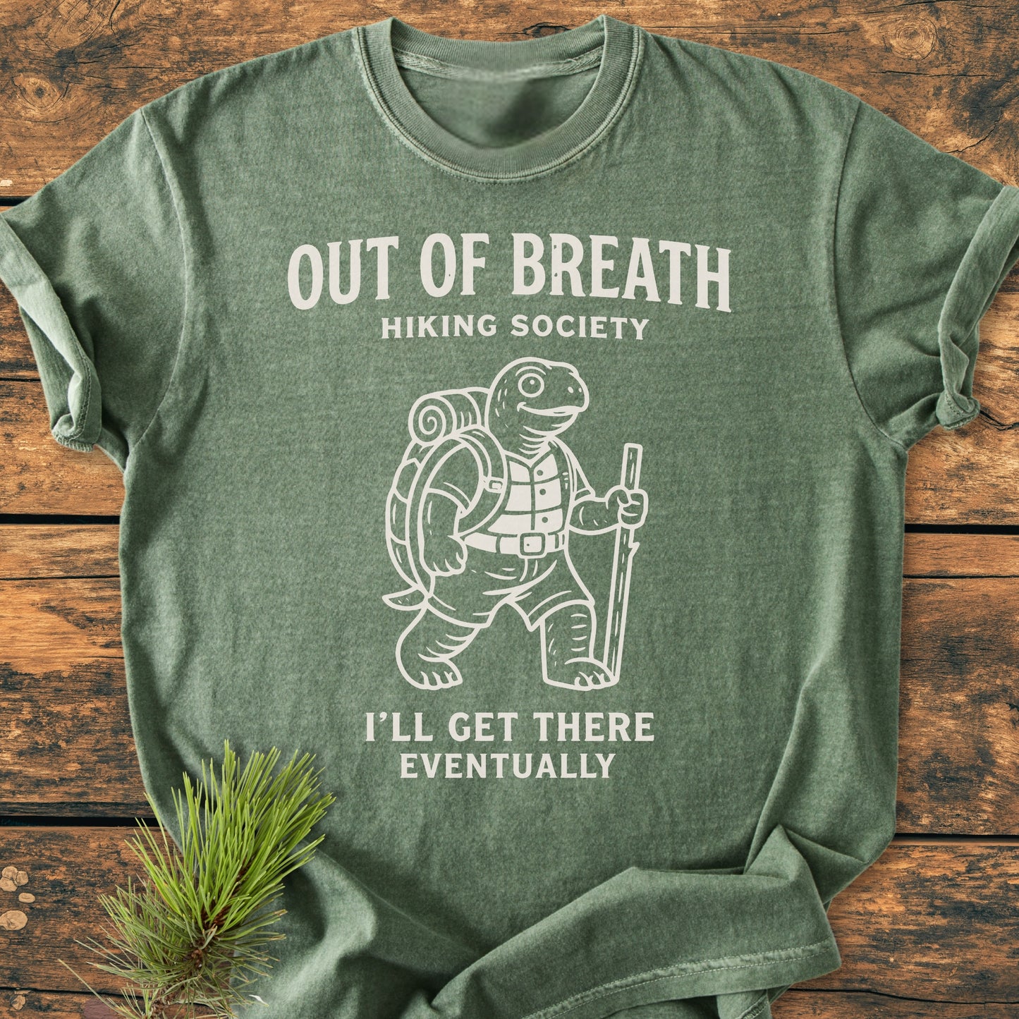 Out Of Breath Turtle - Vintage Wash Tee features a cartoon tortoise with hiking gear and humorous text on a heather-green T-shirt, laid flat on a wood surface, highlighting its relaxed, cozy fit.
