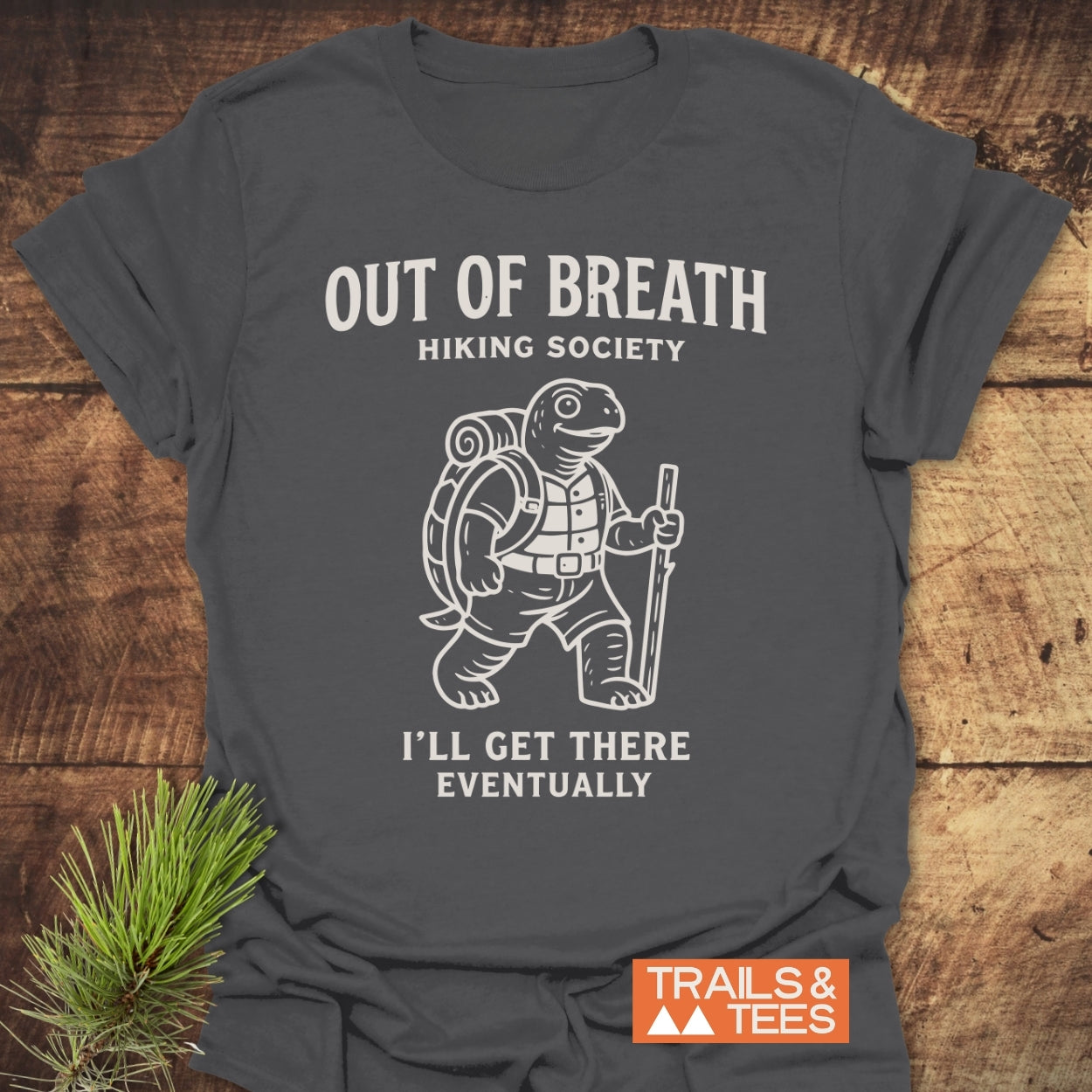 Out Of Breath Turtle T-Shirt featuring a white turtle graphic with hiking gear, surrounded by humorous text, laid on wood; includes “TRAILS & TEES” logo, emphasizing trail-ready comfort and style.