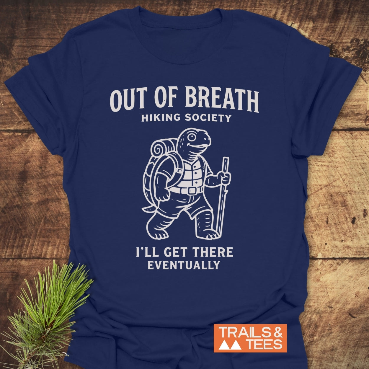 Out Of Breath Turtle T-Shirt featuring a cartoon tortoise with hiking gear and I’LL GET THERE EVENTUALLY slogan, laid on wood with a pine sprig beside it, showcasing outdoor-themed comfort.