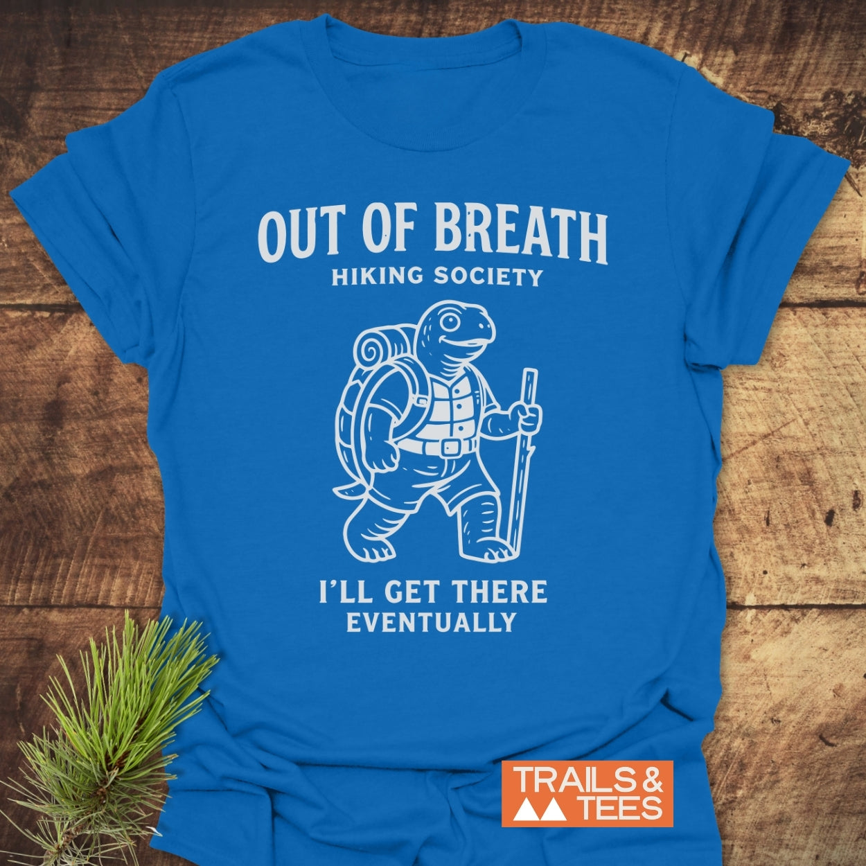 Out Of Breath Turtle T-Shirt featuring a cartoon turtle in hiking gear with the phrase “OUT OF BREATH HIKING SOCIETY” and caption “I’LL GET THERE EVENTUALLY,” laid on a wooden surface.