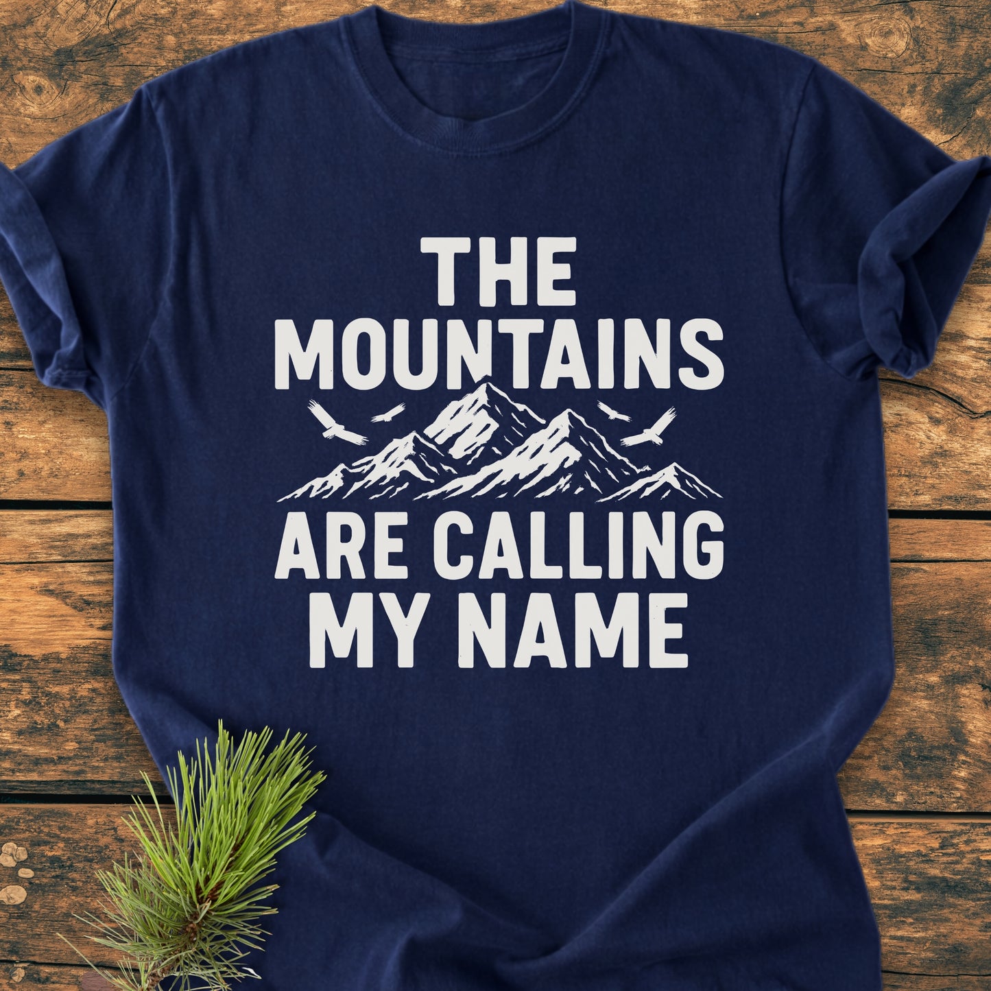 Calling My Name - Vintage Wash Tee: Navy-blue T-shirt with bold white lettering and mountain design, laid on wood surface with pine sprig. Crafted from soft, garment-dyed, 100% ring-spun cotton for comfort.