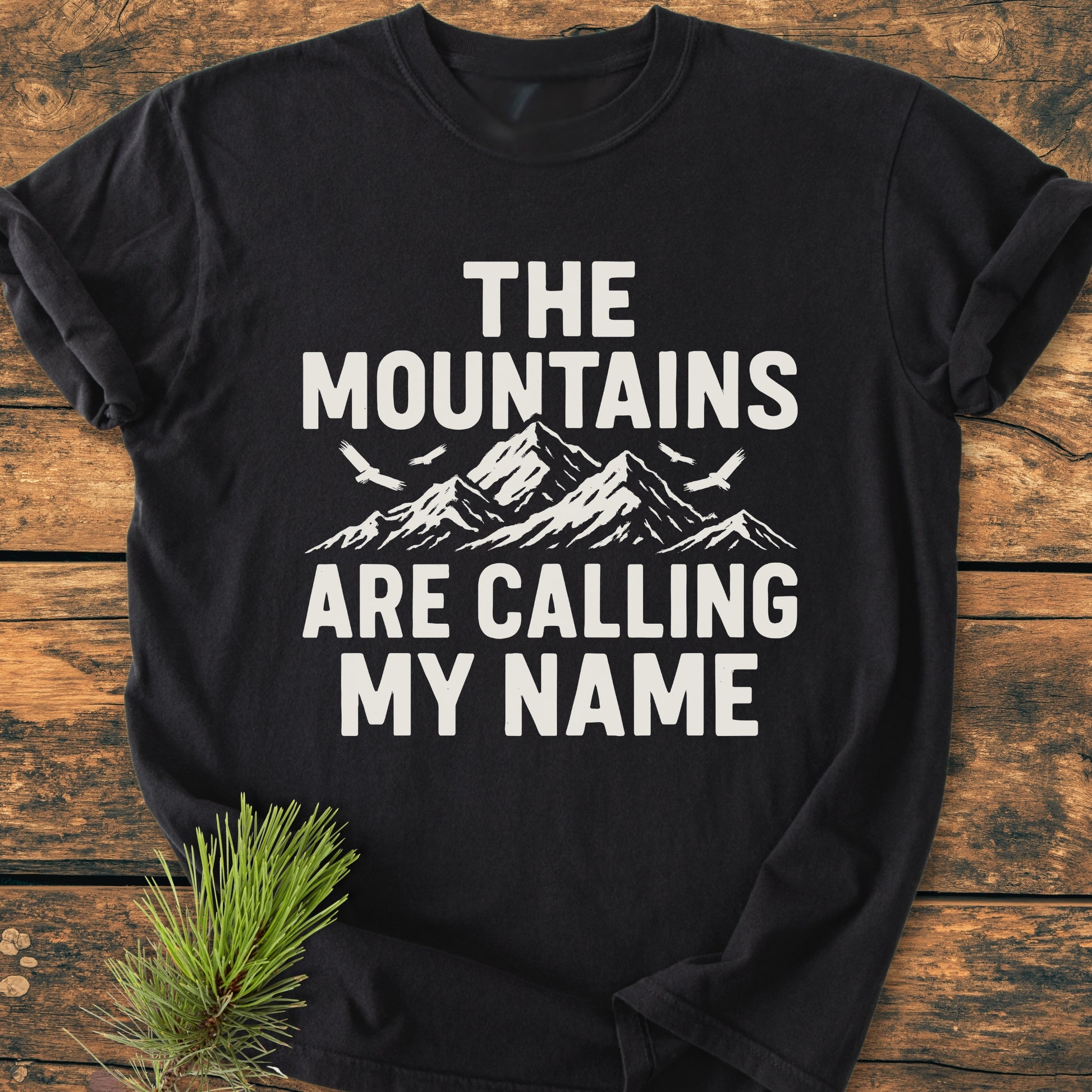 Calling My Name - Vintage Wash Tee features a stylized mountain design with birds and slogan on a black T-shirt, laid on rustic wood with a pine sprig nearby, emphasizing comfort and durability.