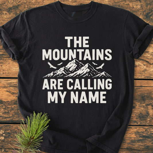 Calling My Name - Vintage Wash Tee features a stylized mountain design with birds and slogan on a black T-shirt, laid on rustic wood with a pine sprig nearby, emphasizing comfort and durability.