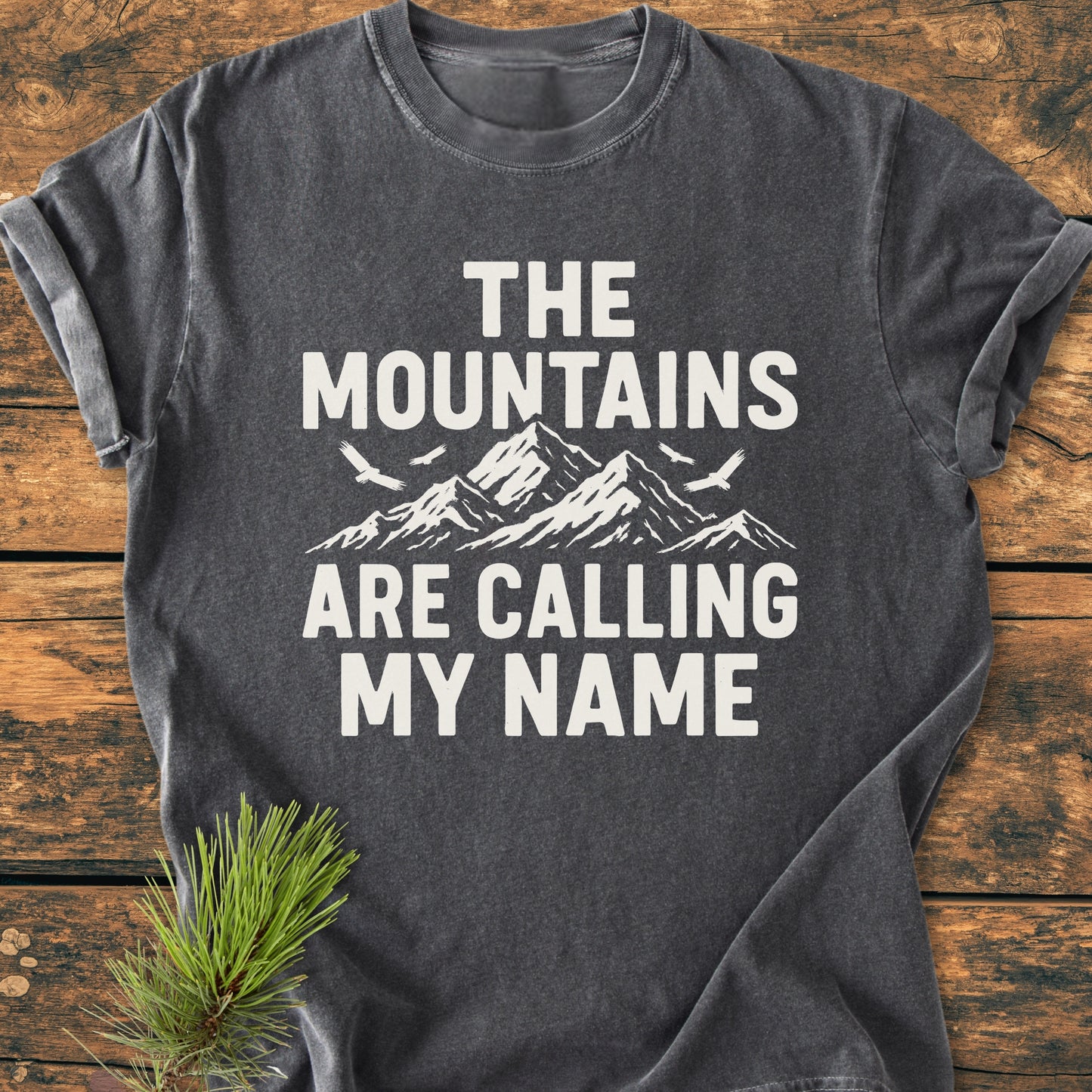 Calling My Name - Vintage Wash Tee in dark gray with bold white text and a mountain graphic. Laid flat on wood, complemented by a pine branch, emphasizing its outdoor theme.