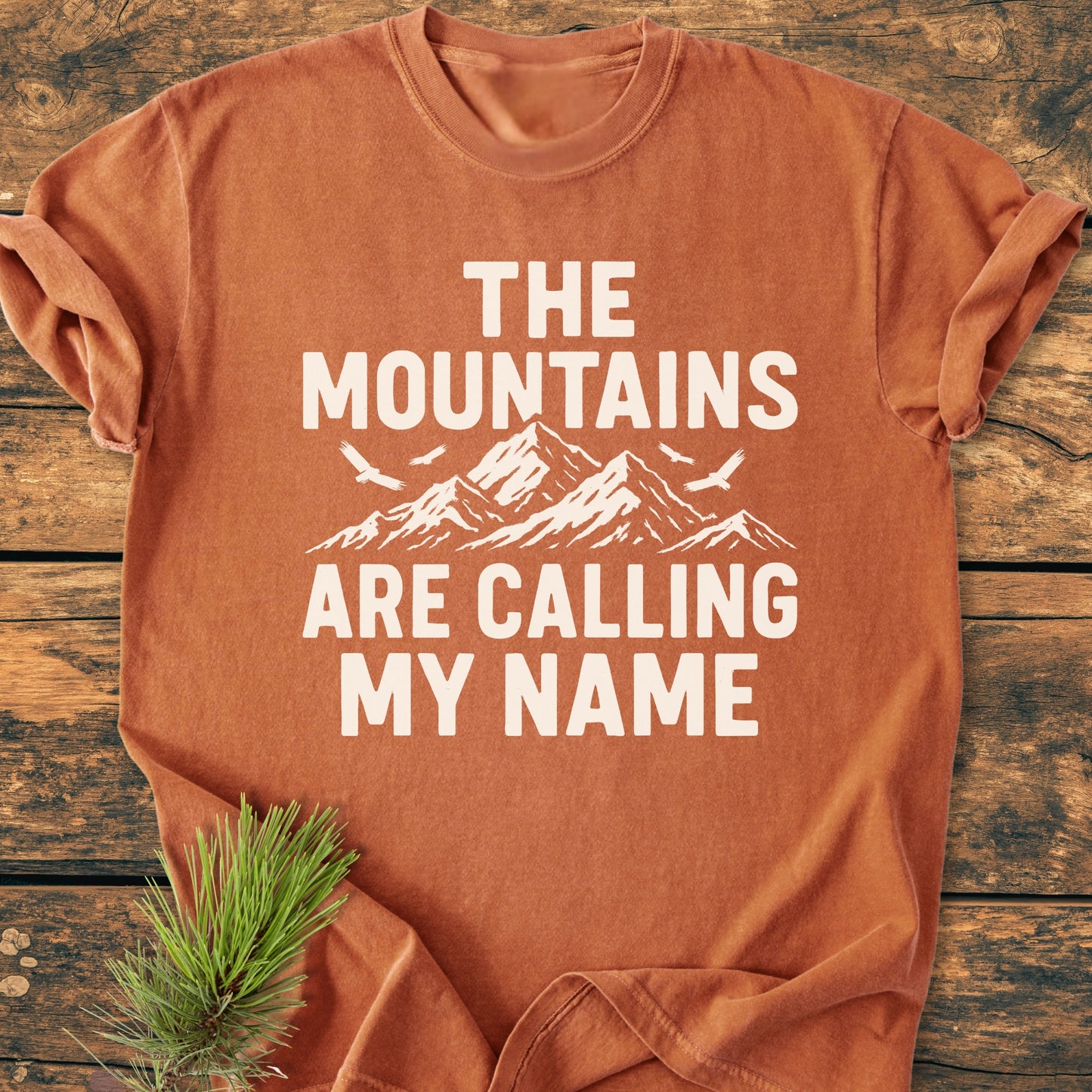 Calling My Name - Vintage Wash Tee: Rust-orange T-shirt with rolled sleeves, featuring a mountain and bird print, and bold text. Displayed on wooden surface, complemented by a pine sprig.