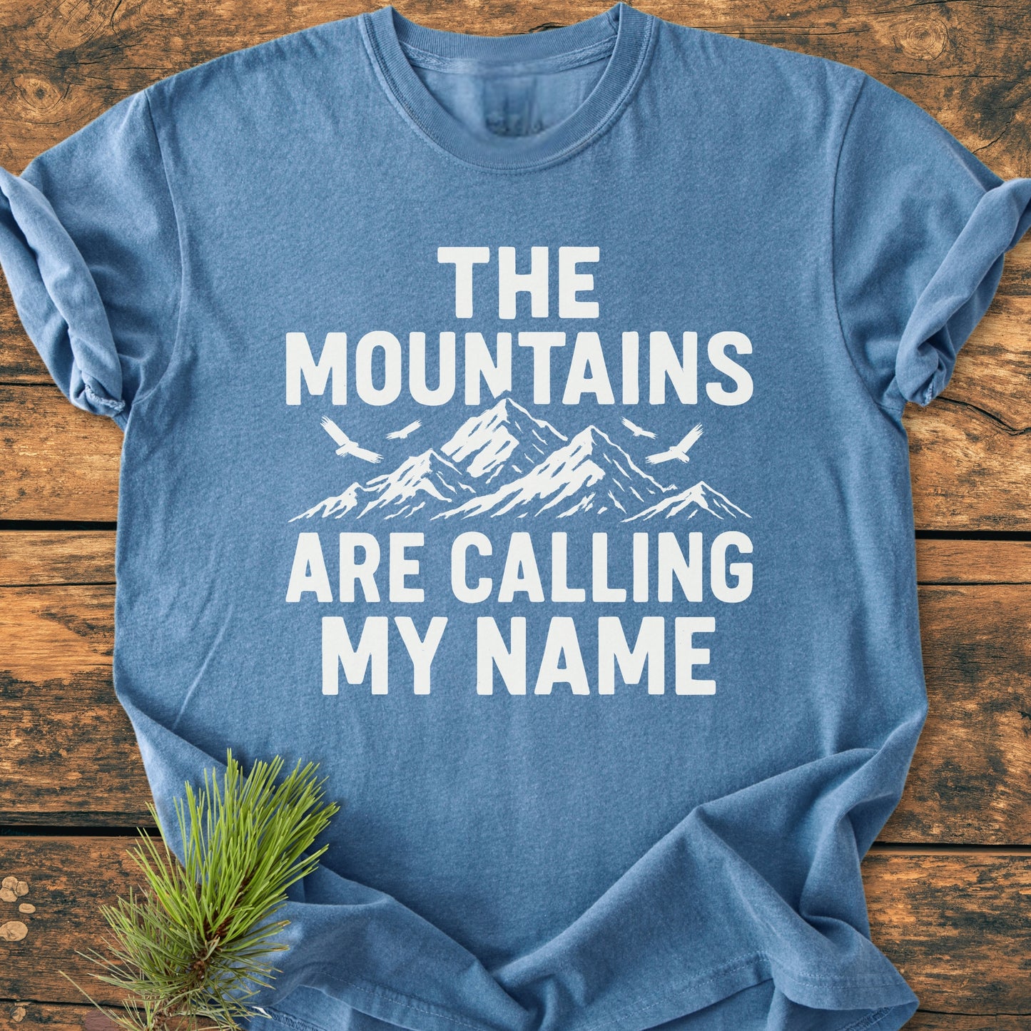 Calling My Name - Vintage Wash Tee featuring a white mountain graphic with birds and text on a heather-blue shirt, styled with a pine sprig on a wooden surface.