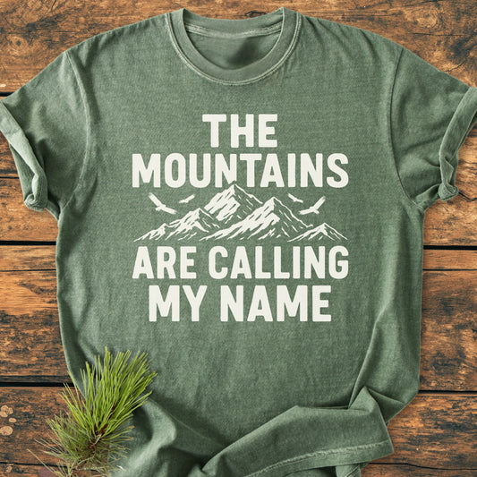 Flat-lay of the Calling My Name - Vintage Wash Tee with stylized mountain graphic and text on a wooden surface, accompanied by a pine branch, highlighting its relaxed fit and durable design.