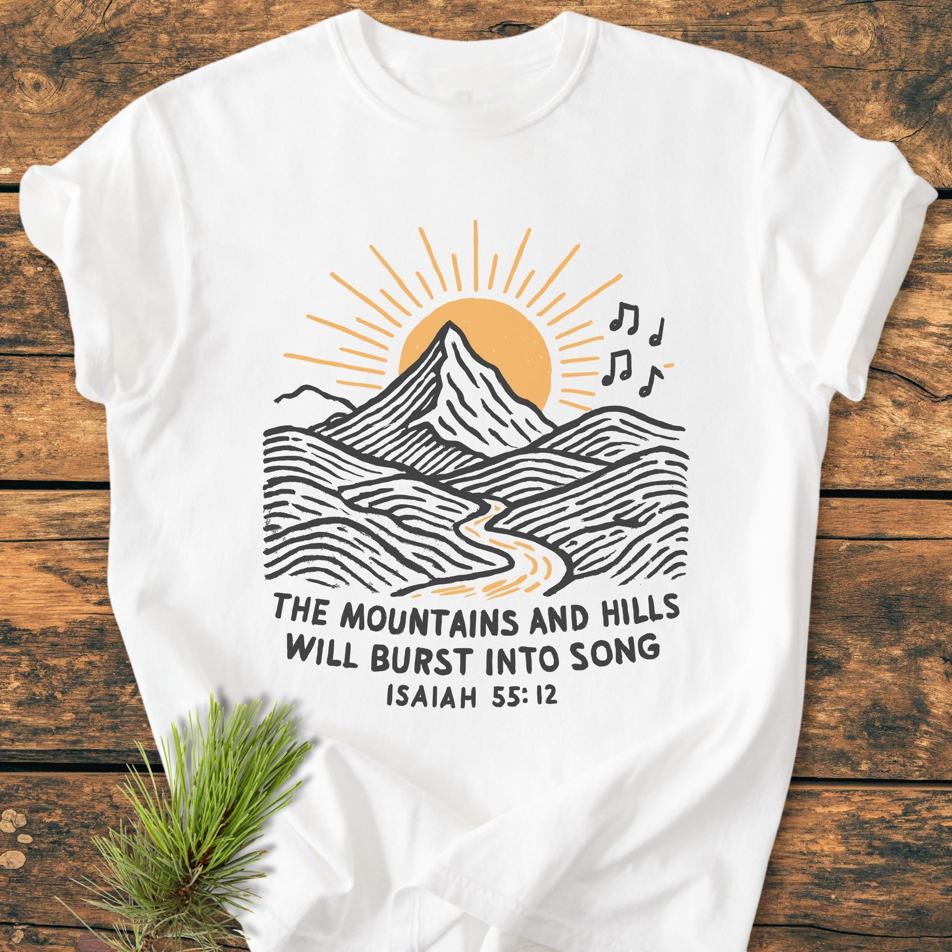 Mountains And Hills - Vintage Wash Tee features a mountain and sun graphic with musical notes and Isaiah 55:12 text, laid on wood with a pine sprig accentuating its rustic charm.